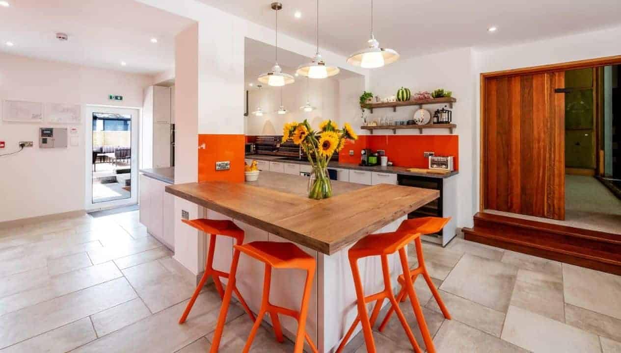 Monnow Valley Studio, Monmouthshire – Modern kitchen with orange-painted accent wall, wooden island and bright orange bar stools.