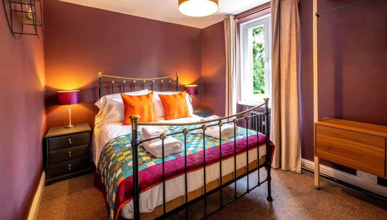 Monnow Valley Studio, Monmouthshire – Bedroom with burgundy walls, metal bed frame, and orange scatter cushions.