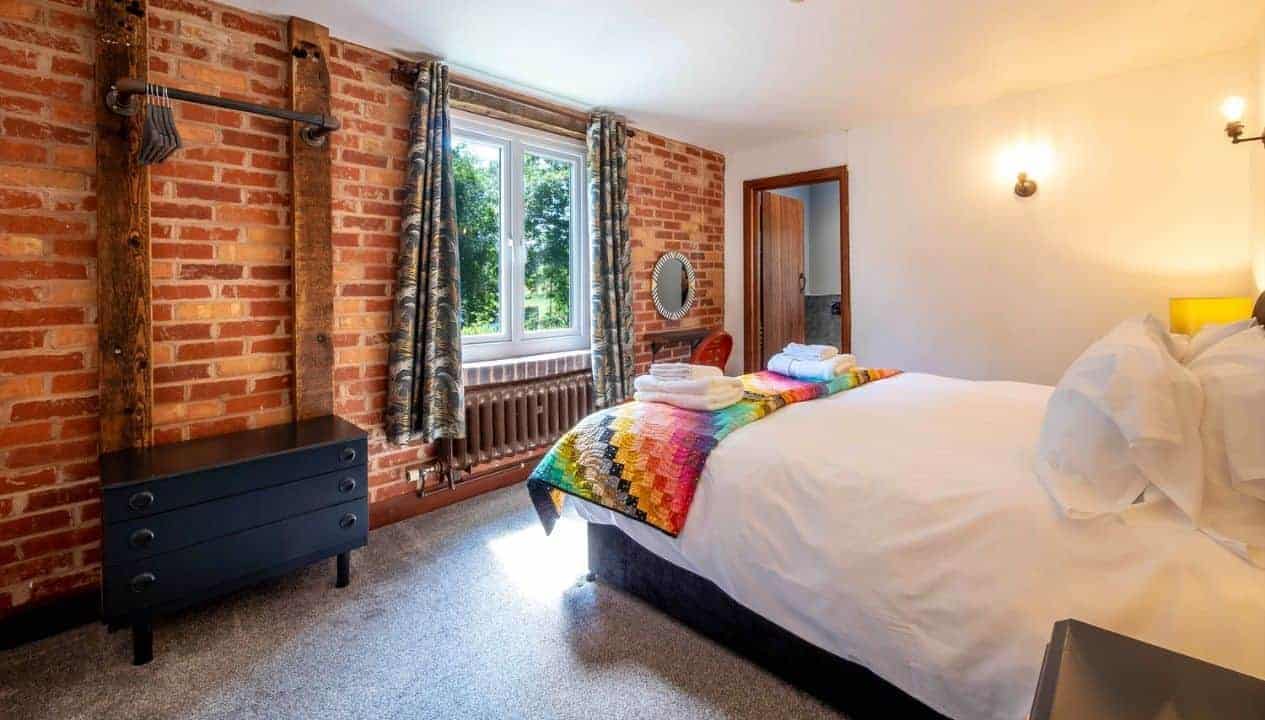 Monnow Valley Studio, Monmouthshire – Bedroom with exposed red brick walls, double bed and patterned throw, large window overlooking grounds.