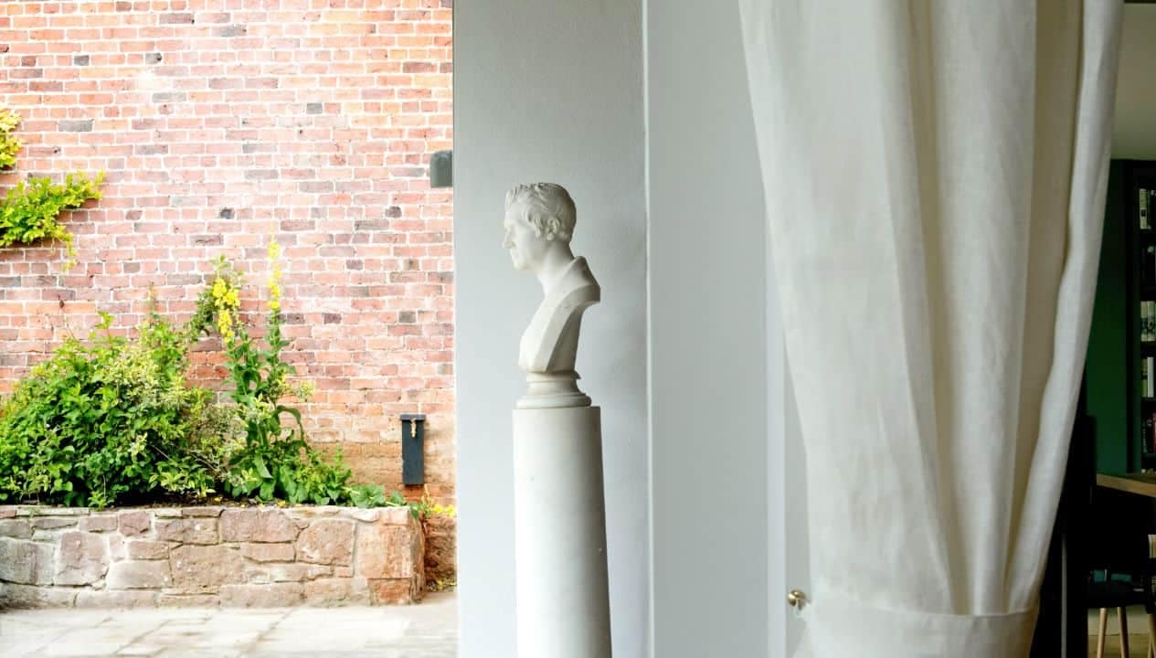 Leuchie Walled Garden – White plaster bust on pedestal beside cream curtains and red brick wall.