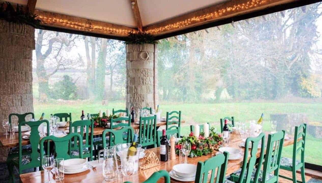 Monnow Valley Studio, Monmouthshire – Covered dining pavilion with green chairs and woodland views through floor-to-ceiling windows.