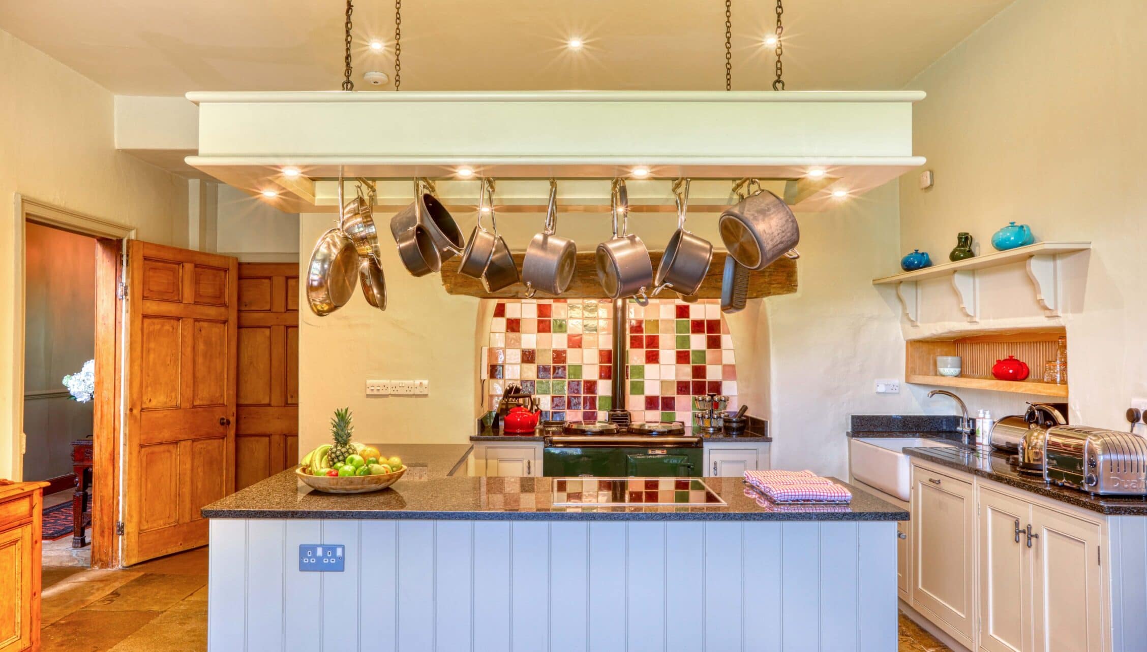 Gitcombe House, Devon – Kitchen with suspended copper pan rack and colourful tiled splashback above Aga cooker.