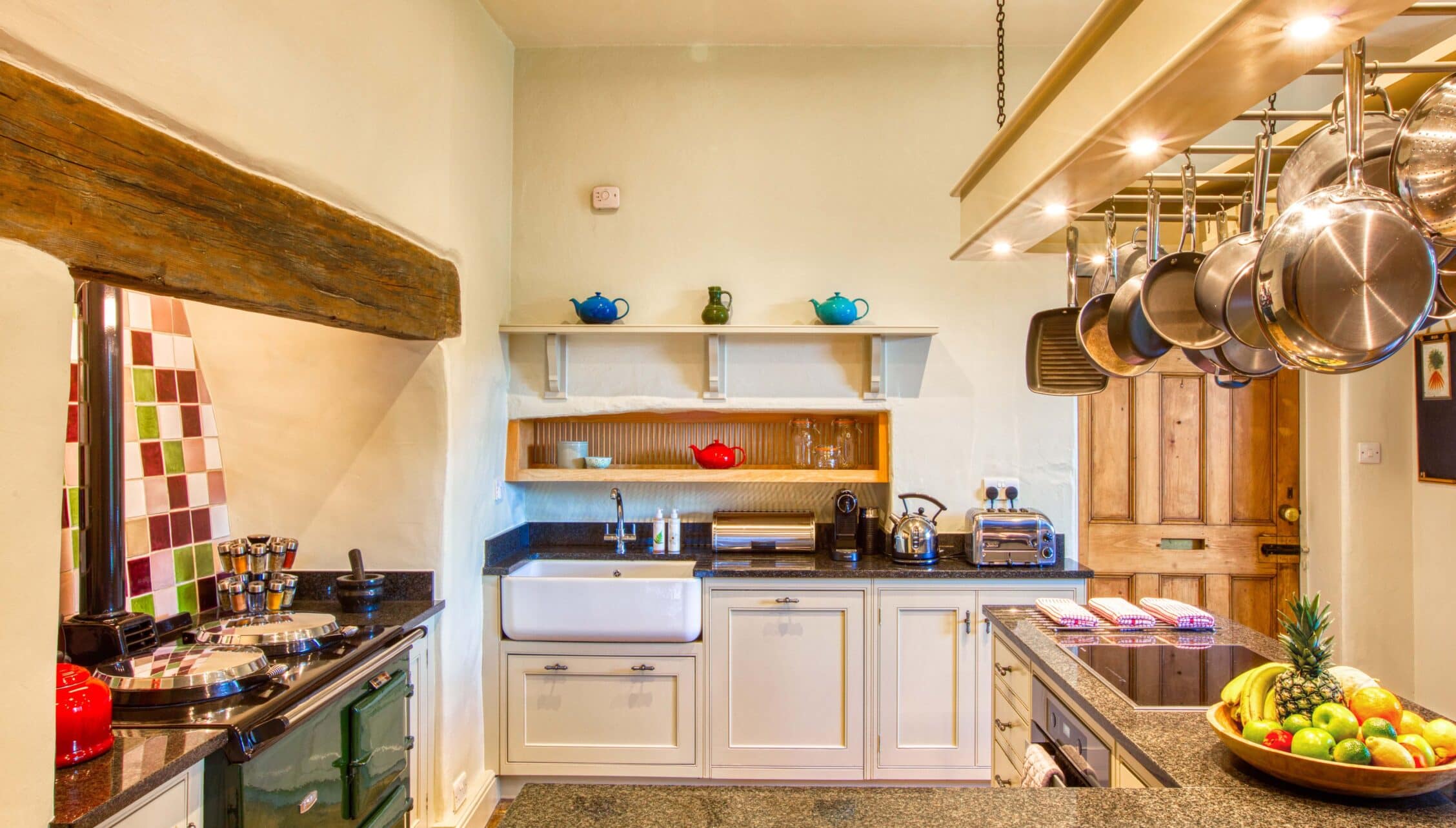 Gitcombe House, Devon – Spacious kitchen with cream cabinetry, exposed wooden beams, and hanging copper cookware.
