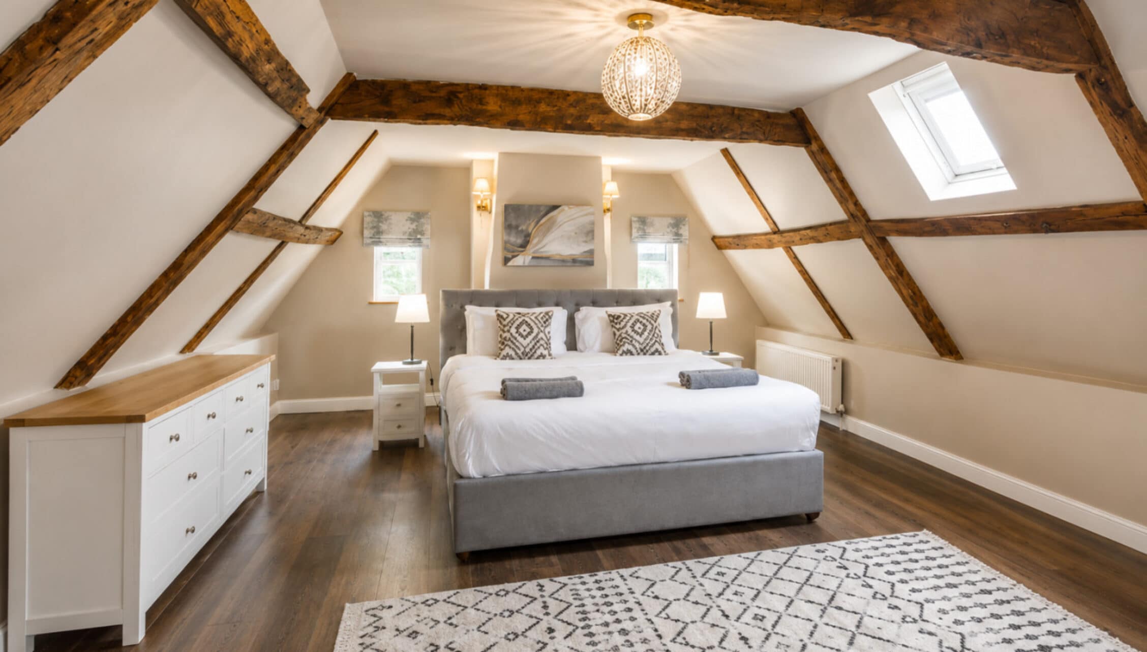 Catton Old Hall, Norfolk – Attic bedroom with exposed wooden beams, grey upholstered bed, white storage dresser.