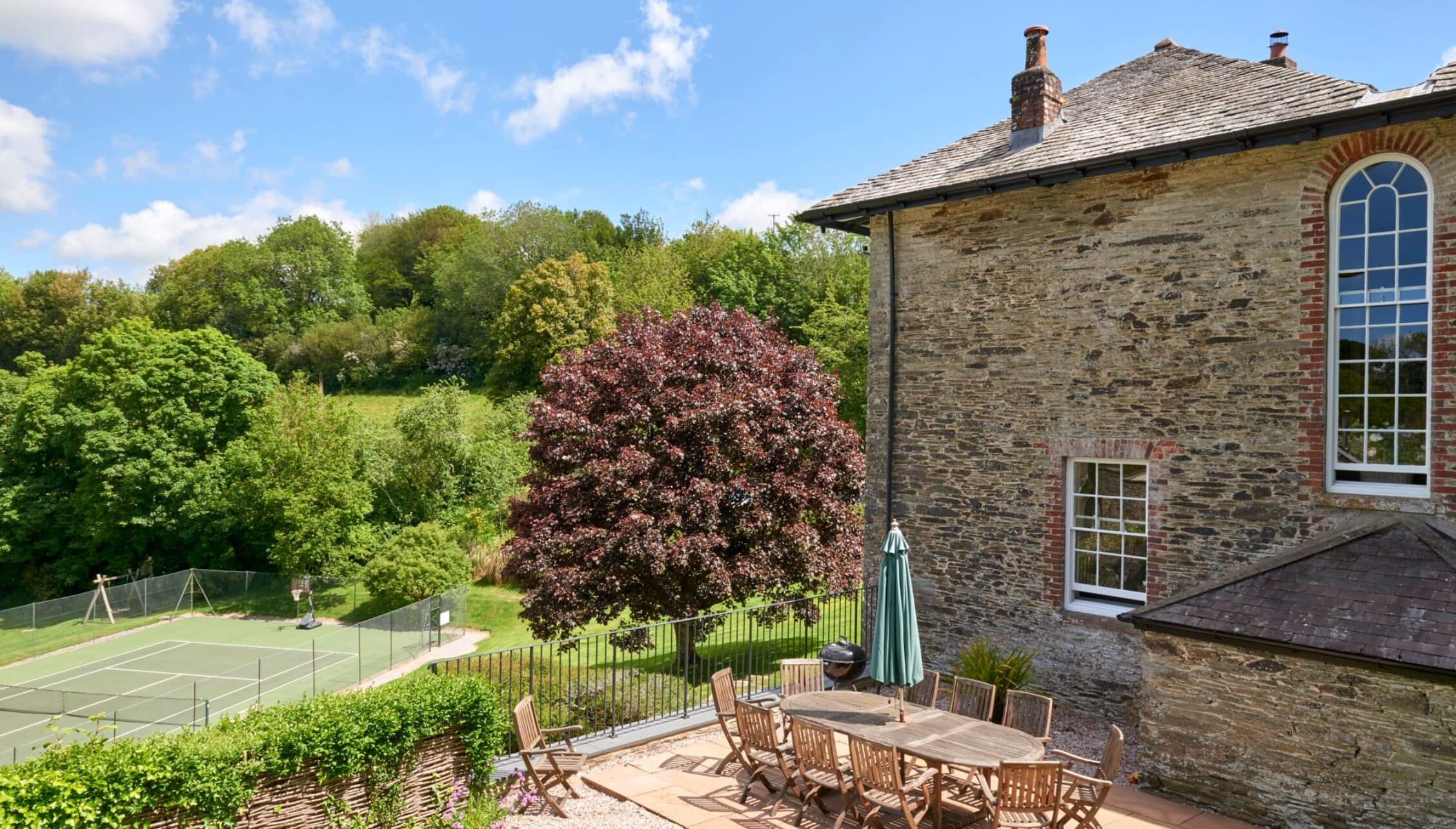 Gitcombe House, Devon – Stone country house with arched windows, patio seating, and tennis court beyond manicured gardens.
