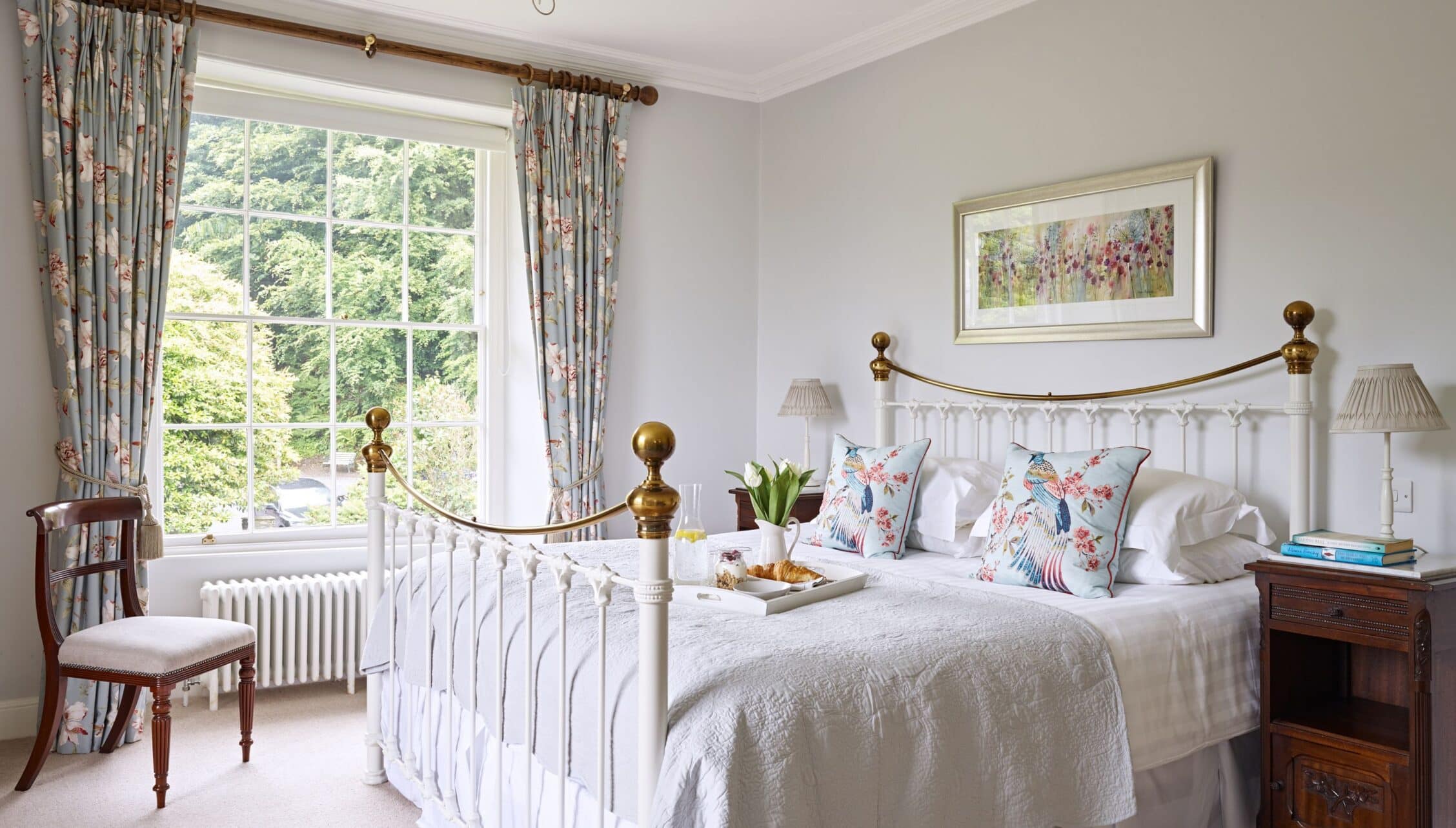 Gitcombe House, Devon – Bedroom with white brass bed frame, floral curtains and garden views through sash window.