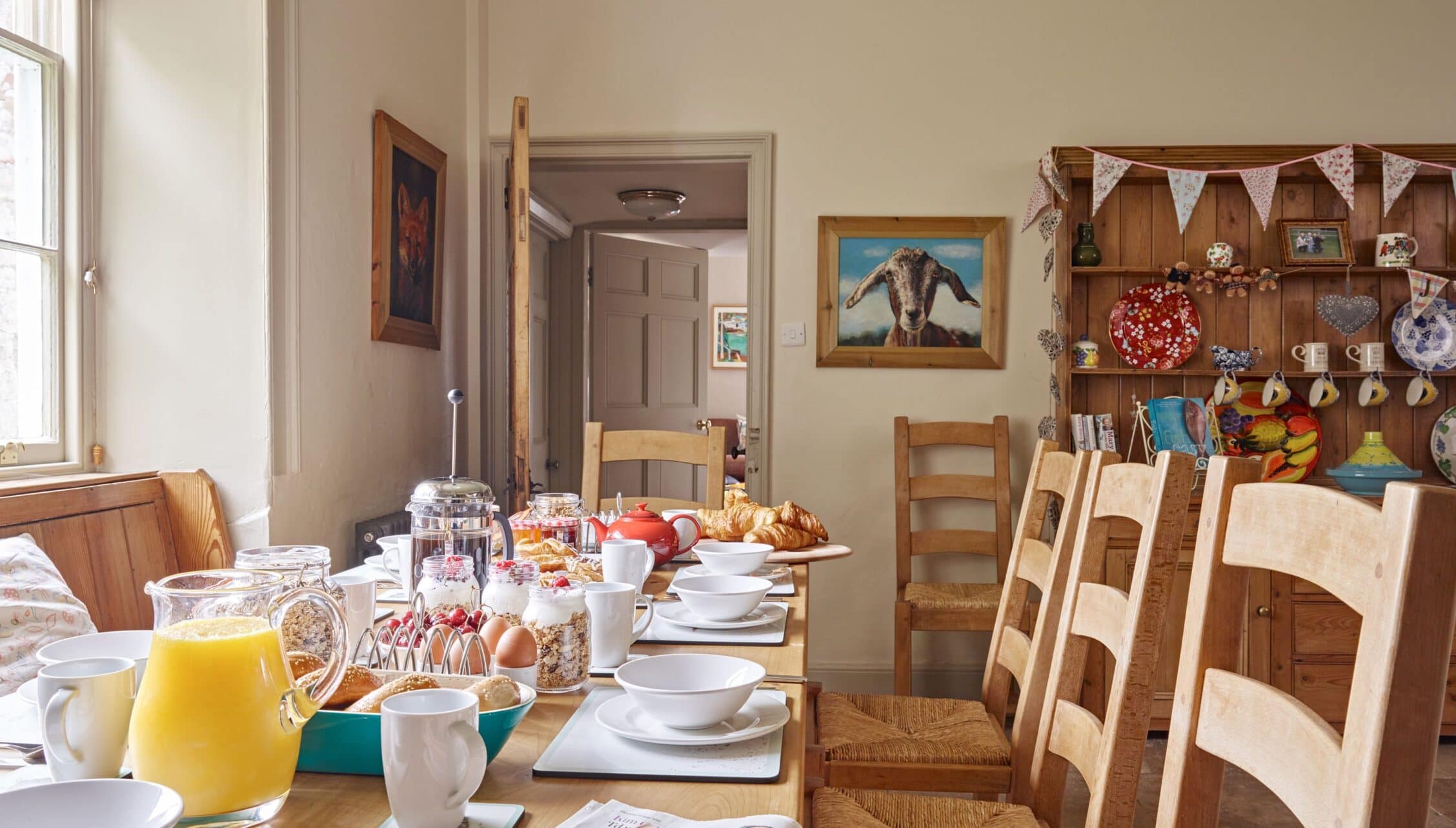 Gitcombe House, Devon – Dining room with wooden chairs, set breakfast table, and open shelving displaying ceramics and decorations.