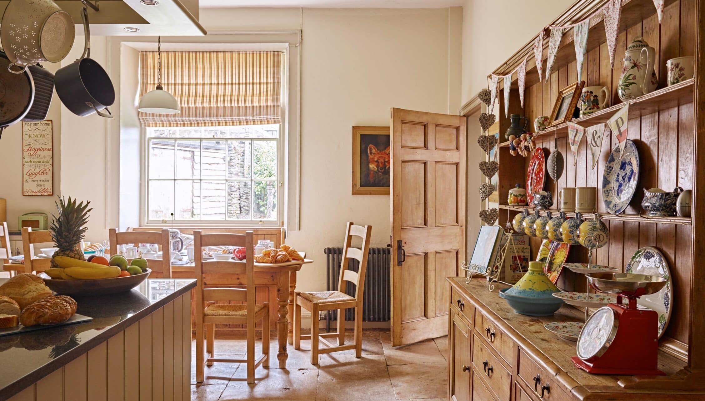 Gitcombe House, Devon – Country kitchen with wooden dresser, striped Roman blind, and hung cookware.