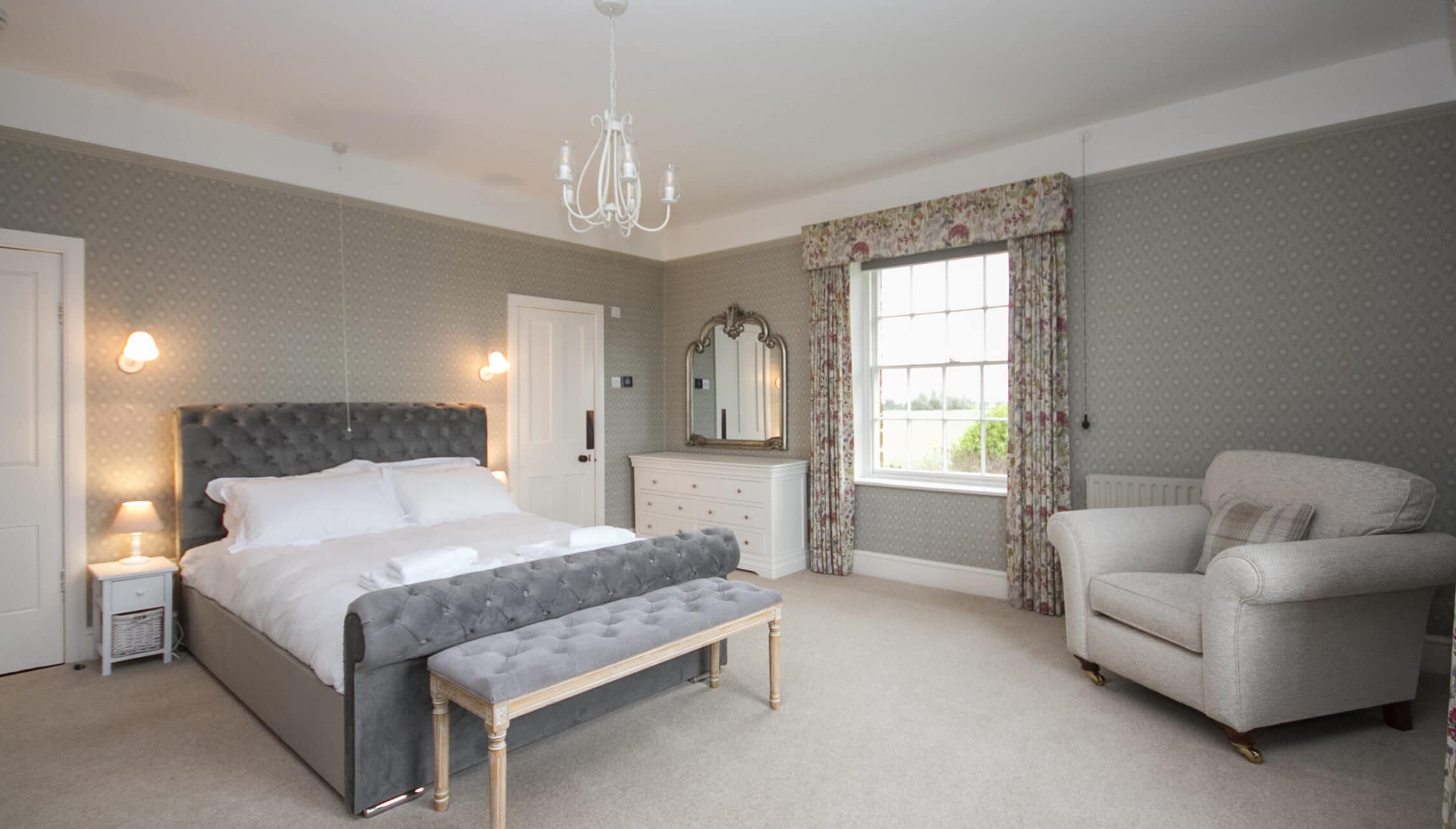 Greens Norton House, Northamptonshire — Spacious master bedroom with grey upholstered bed, floral curtains, and seating area.