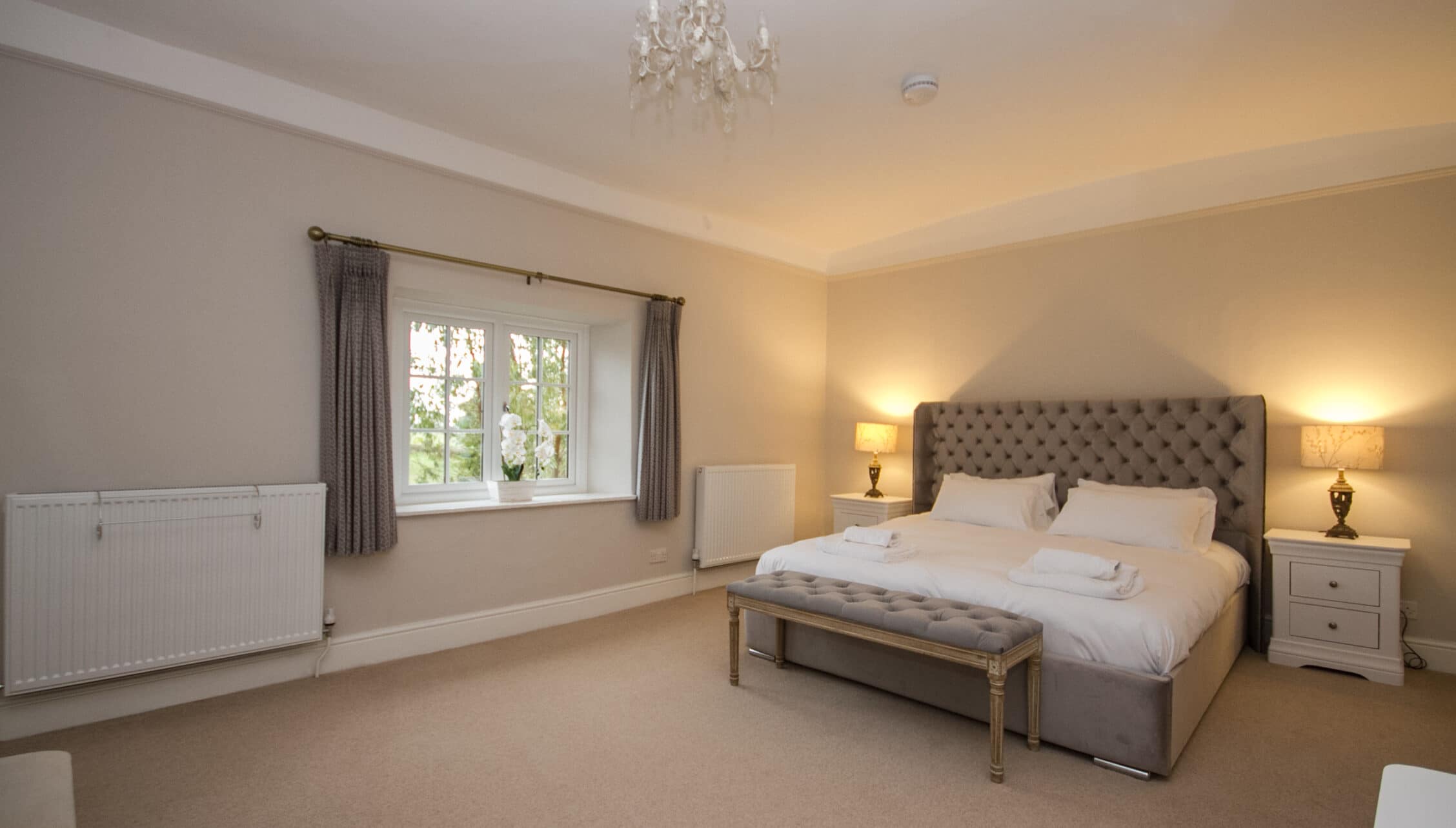 Greens Norton House, Northamptonshire — Spacious master bedroom with upholstered headboard, crystal chandelier, and garden views.