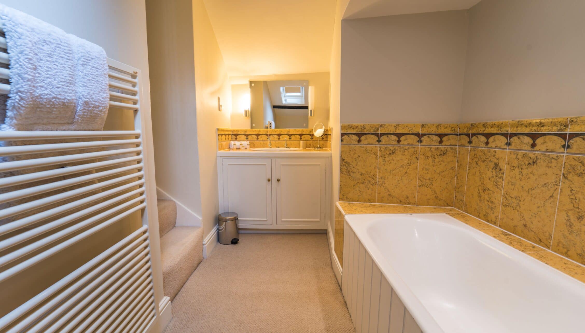 Gitcombe House, Devon – Bathroom with gold-tiled walls, white bath, and heated towel rail.