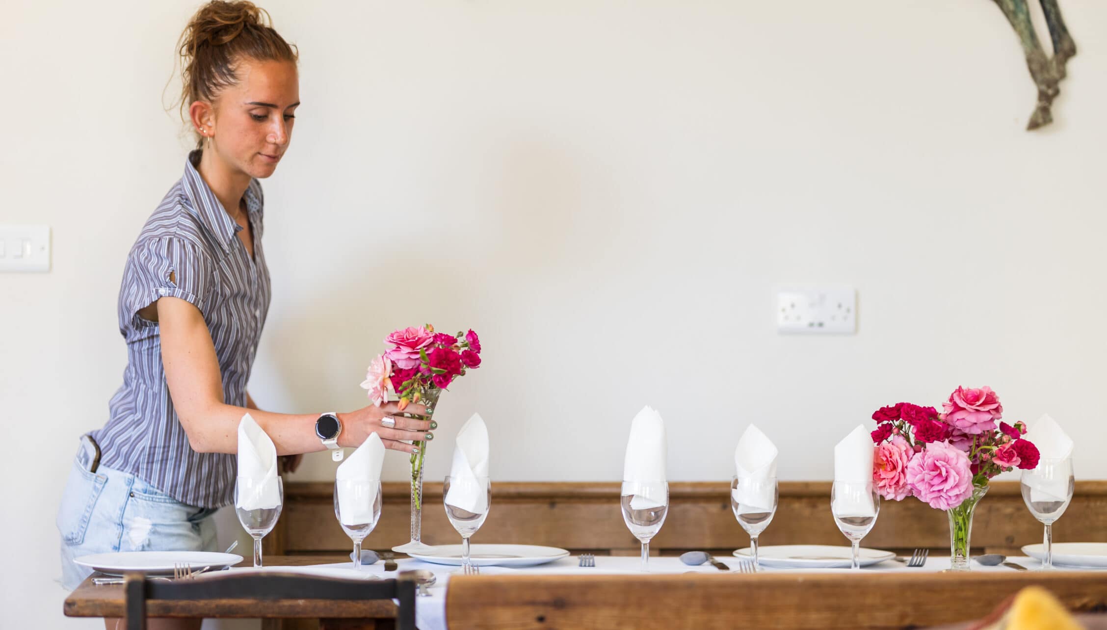 Upper Court, Gloucestershire – Woman setting a dining table with pink and red carnation flowers and white napkins.