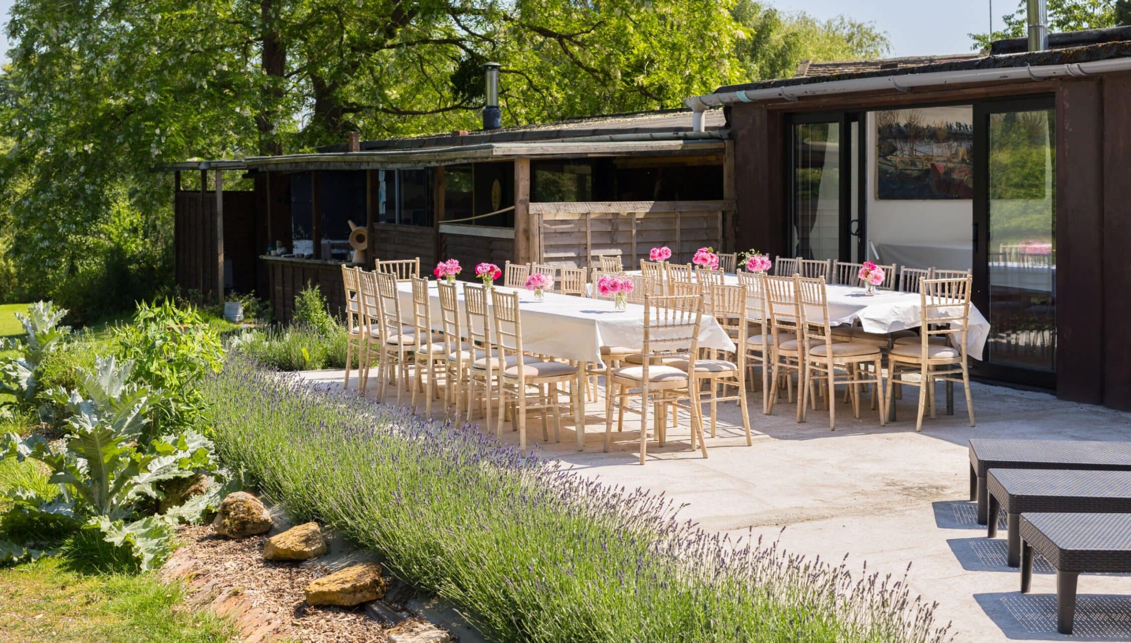 Upper Court, Gloucestershire – Outdoor dining terrace with wooden chairs, pink flowers, and modern pavilion beneath mature trees.