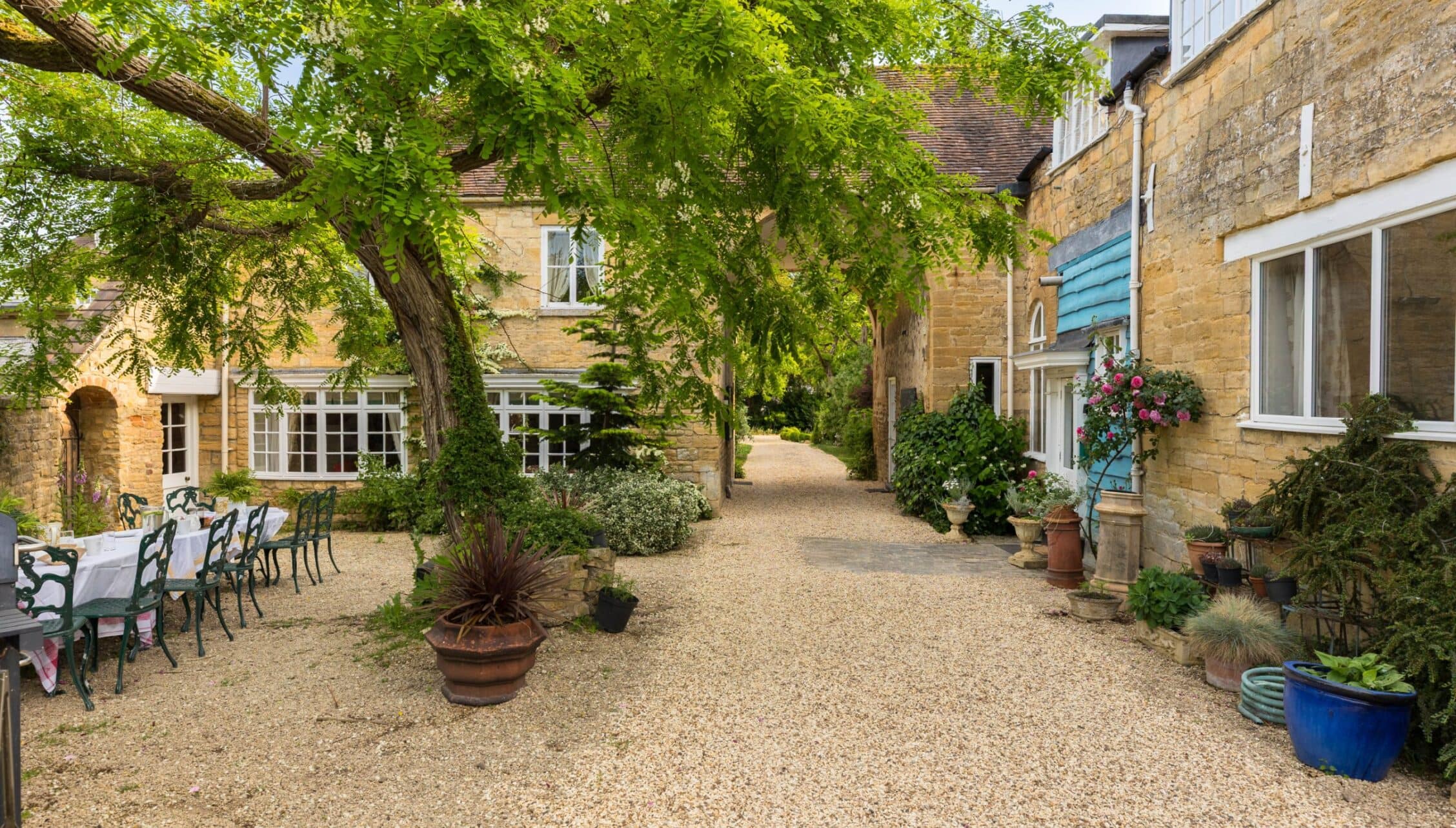 Upper Court, Gloucestershire – Gravel courtyard with stone buildings, mature trees, and potted plants.