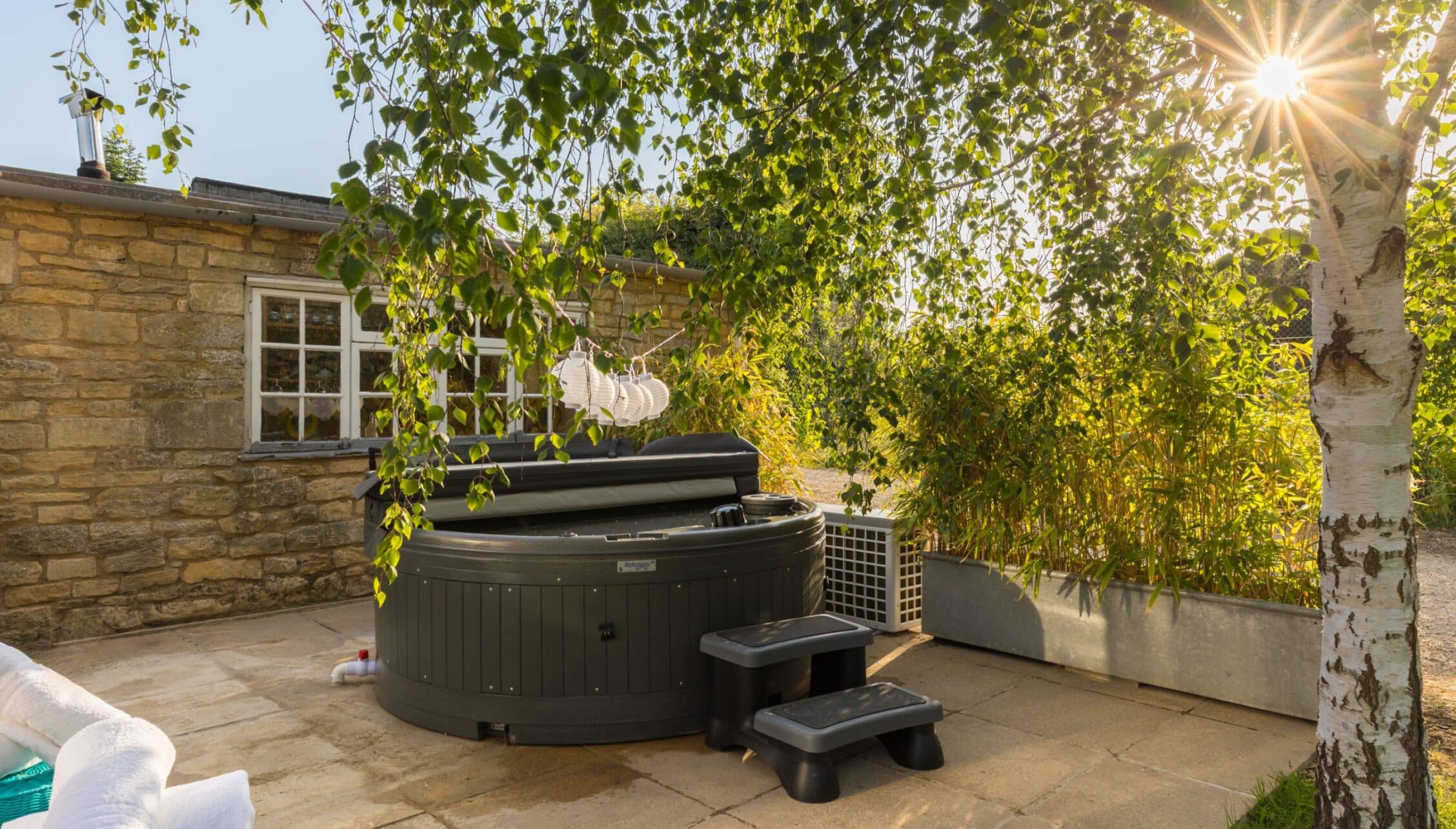 Upper Court, Gloucestershire – Hot tub on stone patio beside stone cottage with birch tree and climbing ivy.