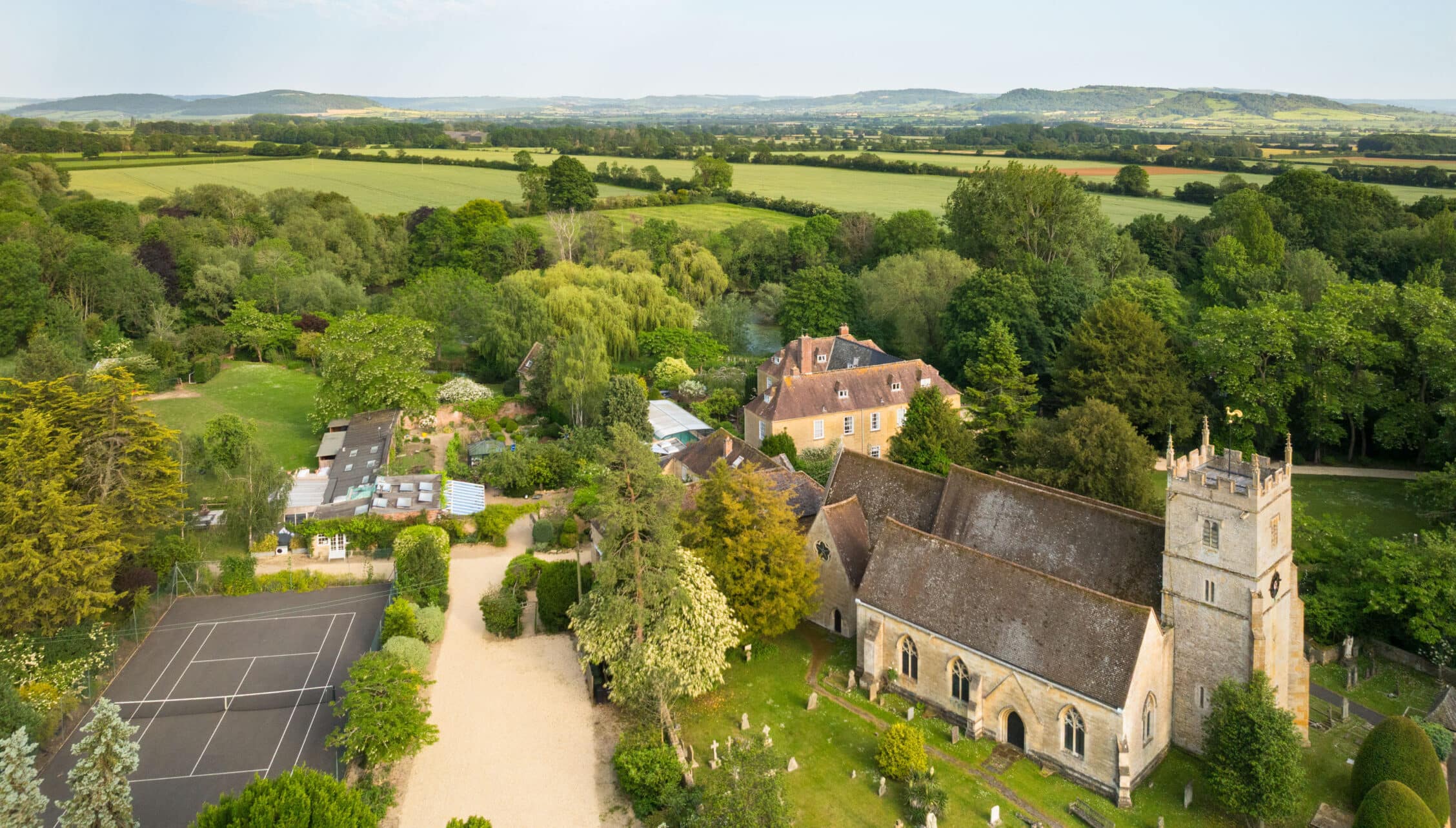 Upper Court, Gloucestershire – Aerial view of stone country house, church, and manicured grounds overlooking rolling farmland.