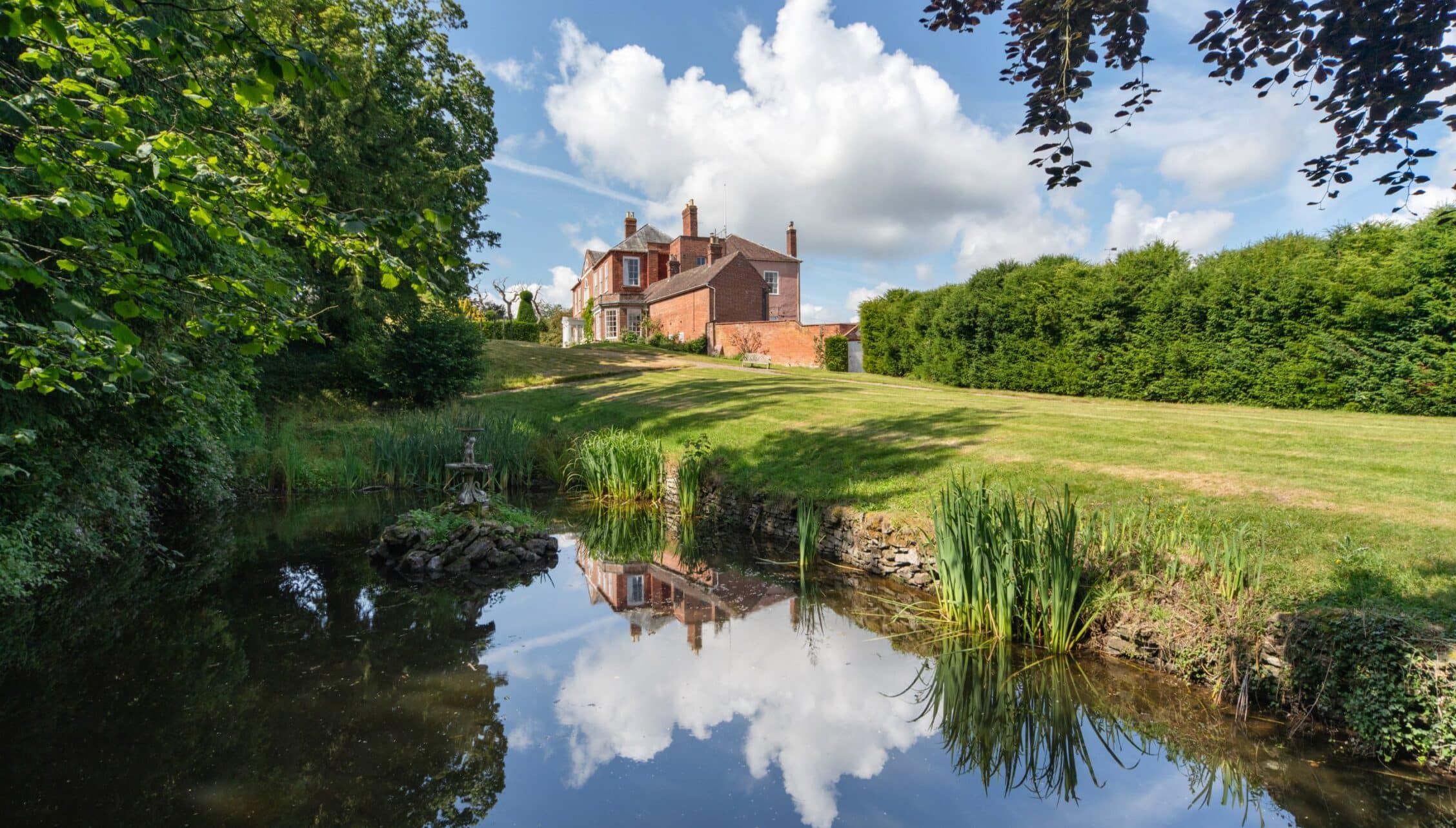 Barton Court, Herefordshire – Red brick country house with pond and manicured lawn bordered by hedges.