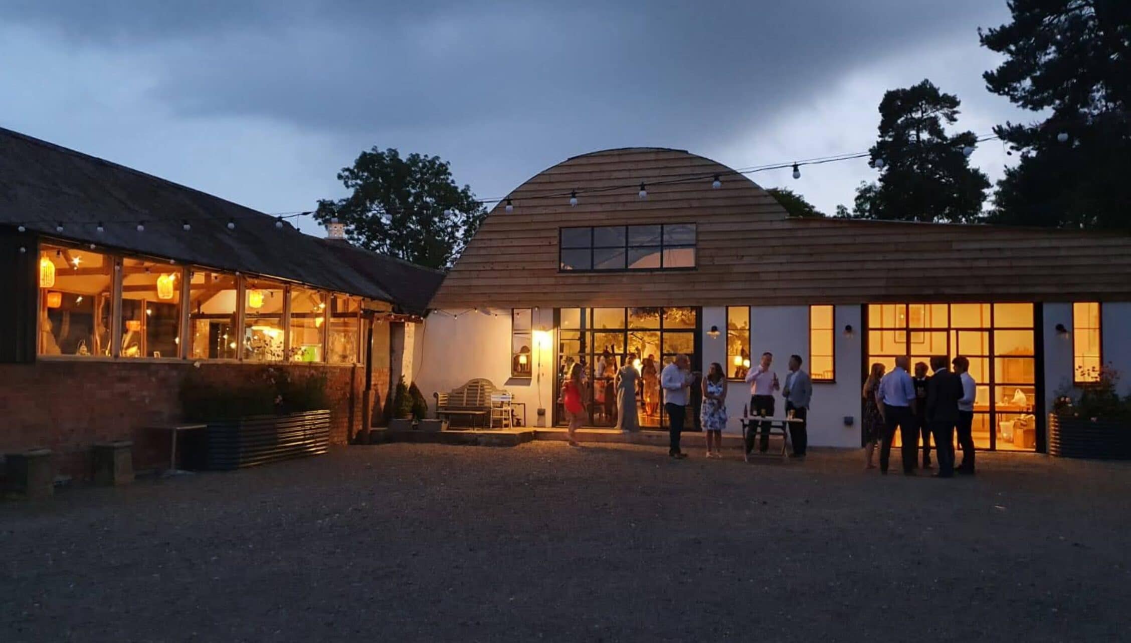 Barton Court, Herefordshire – Modern curved timber-clad building with large windows illuminated at dusk, guests gathered in courtyard.
