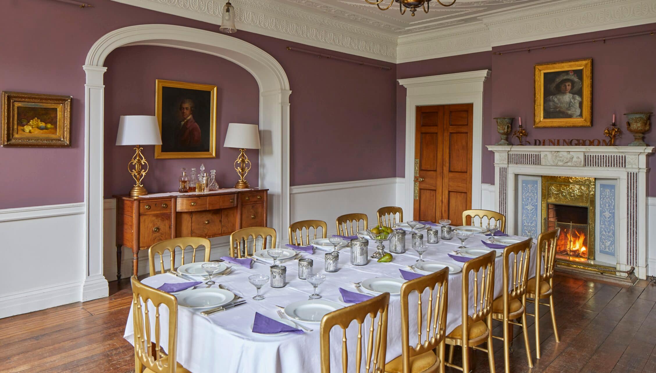 Barton Court, Herefordshire – Formal dining room with purple walls, fireplace and wooden chairs set for dinner.