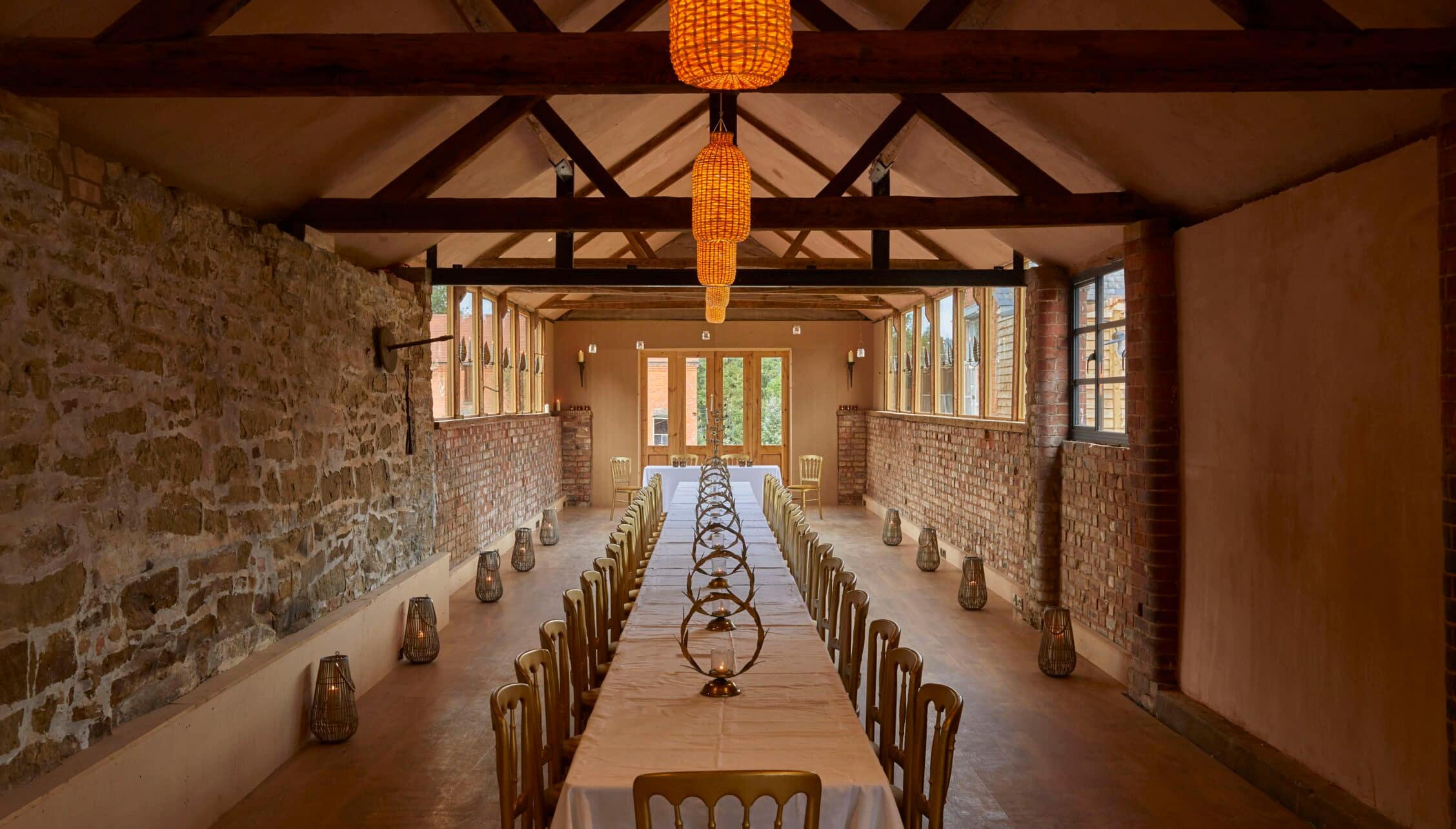Barton Court, Herefordshire – Long stone barn dining room with exposed beams, wicker pendant lights and laid table.