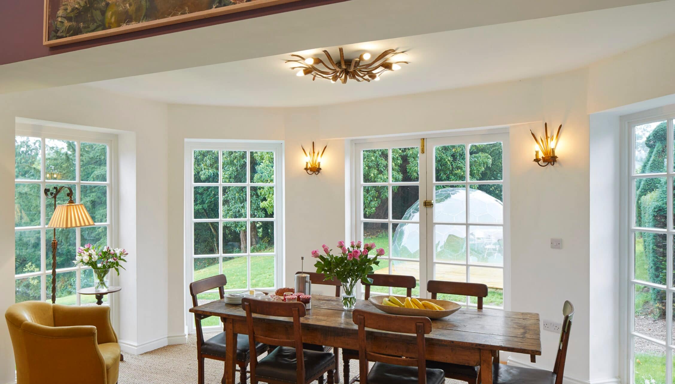 Barton Court, Herefordshire – Dining room with wooden table, brass wall lights and extensive garden views through multi-pane windows.
