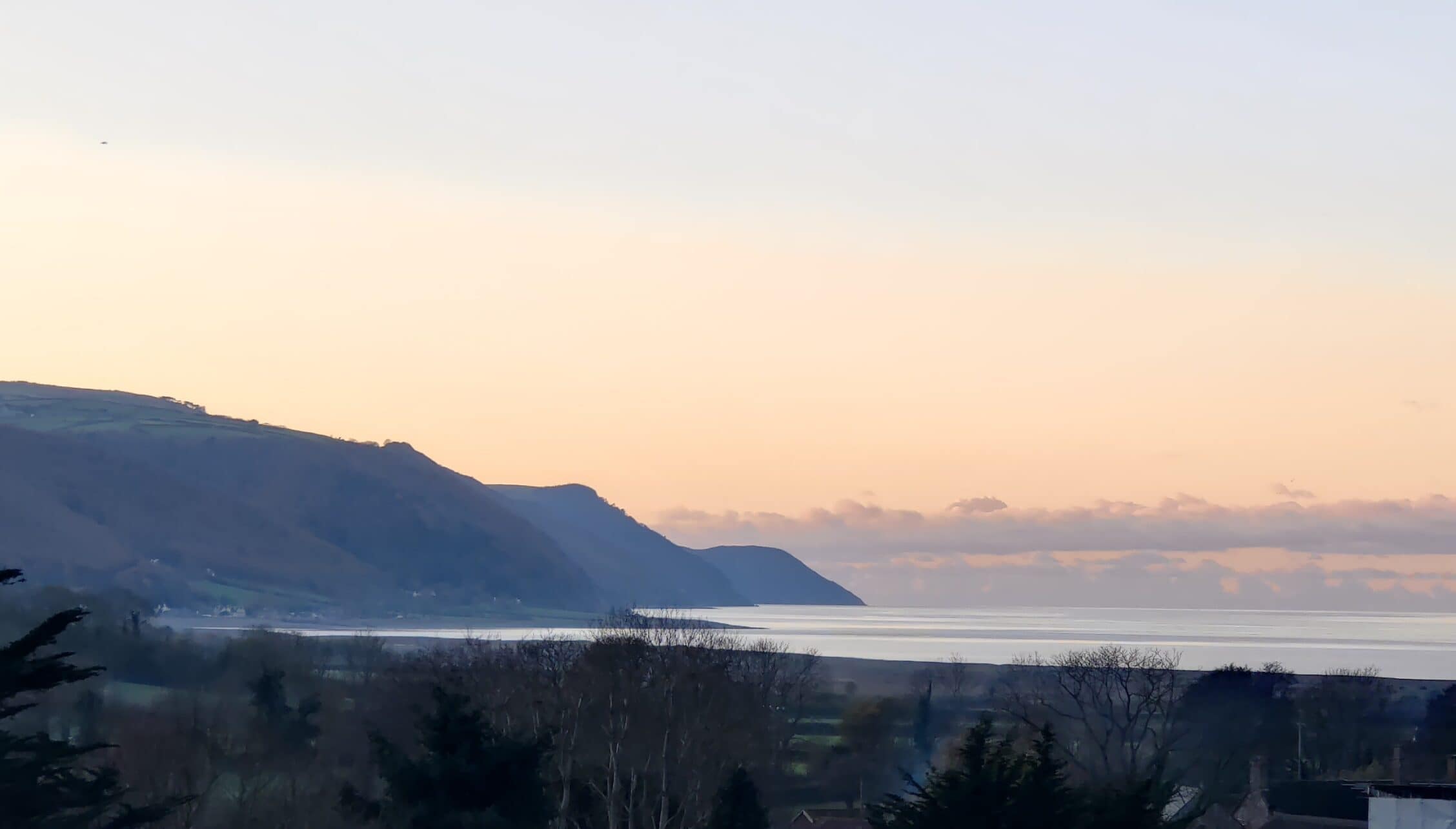 Bossington Hall, Somerset – Coastal sea view at dusk with wooded hillside and distant cliffs.