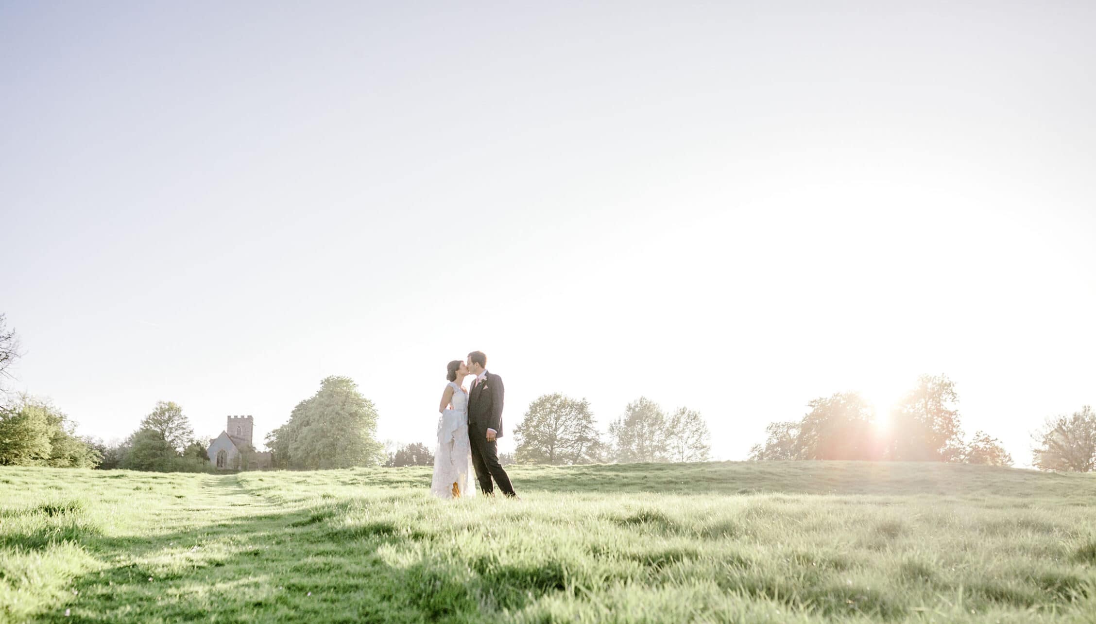Boxted Hall, Suffolk – Bride and groom kissing in open parkland with church and tree-lined horizon.