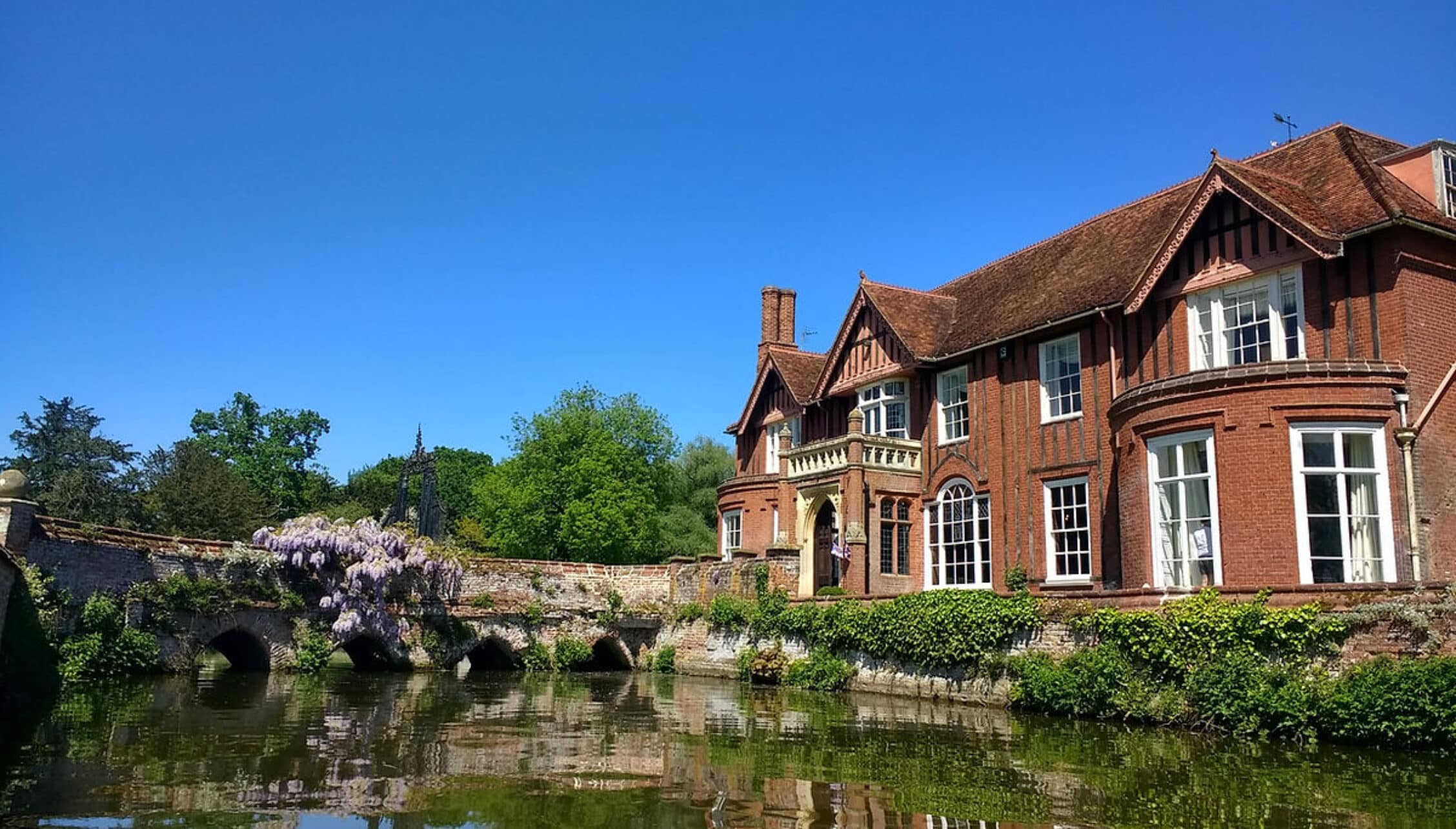 Boxted Hall, Suffolk – Victorian brick manor house with tower reflected in river, stone bridge and wisteria visible.