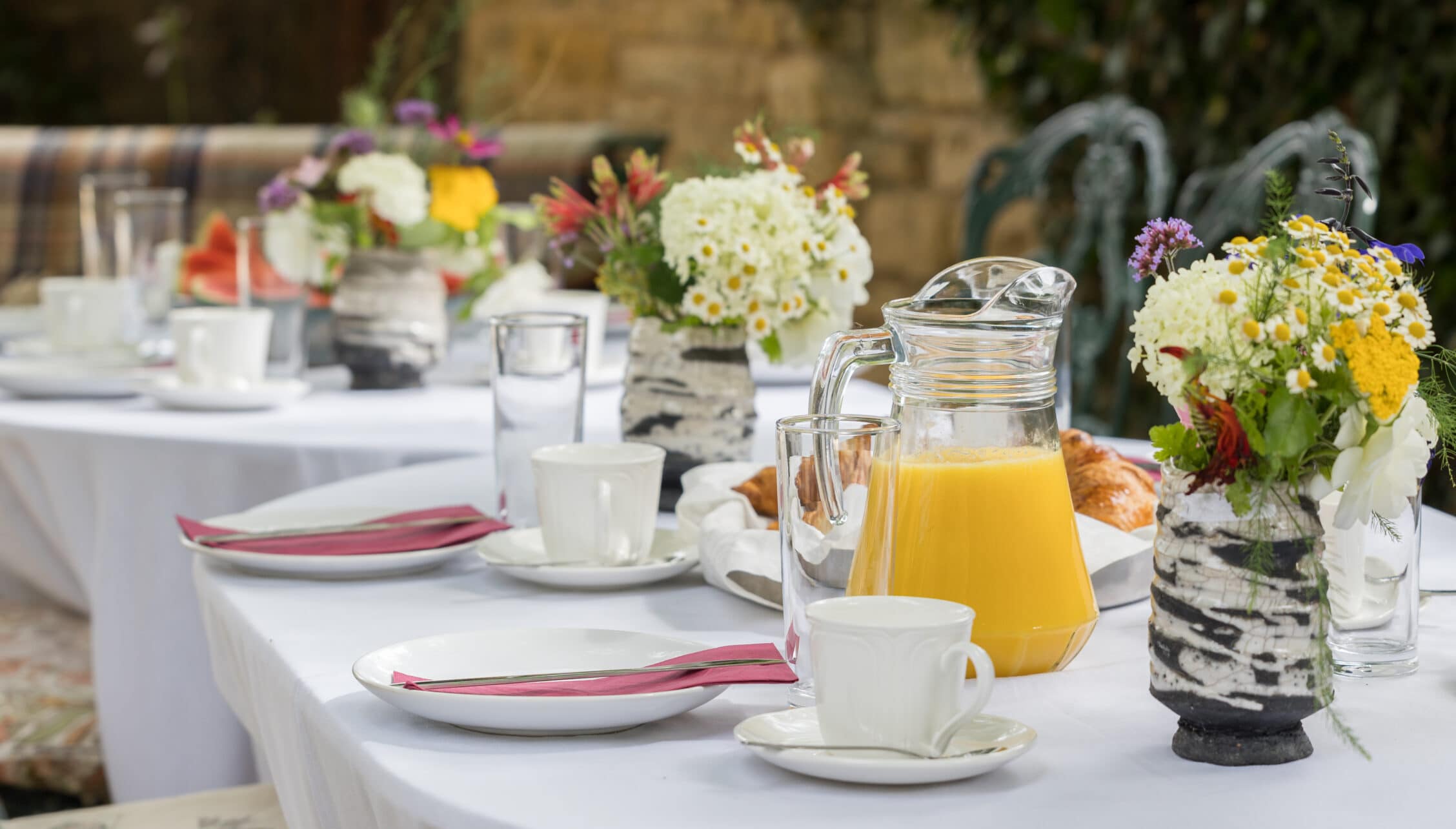 Courtyard Cottages, Gloucestershire – Outdoor dining table set with white linens, pink napkins, floral centrepieces and juice pitcher.