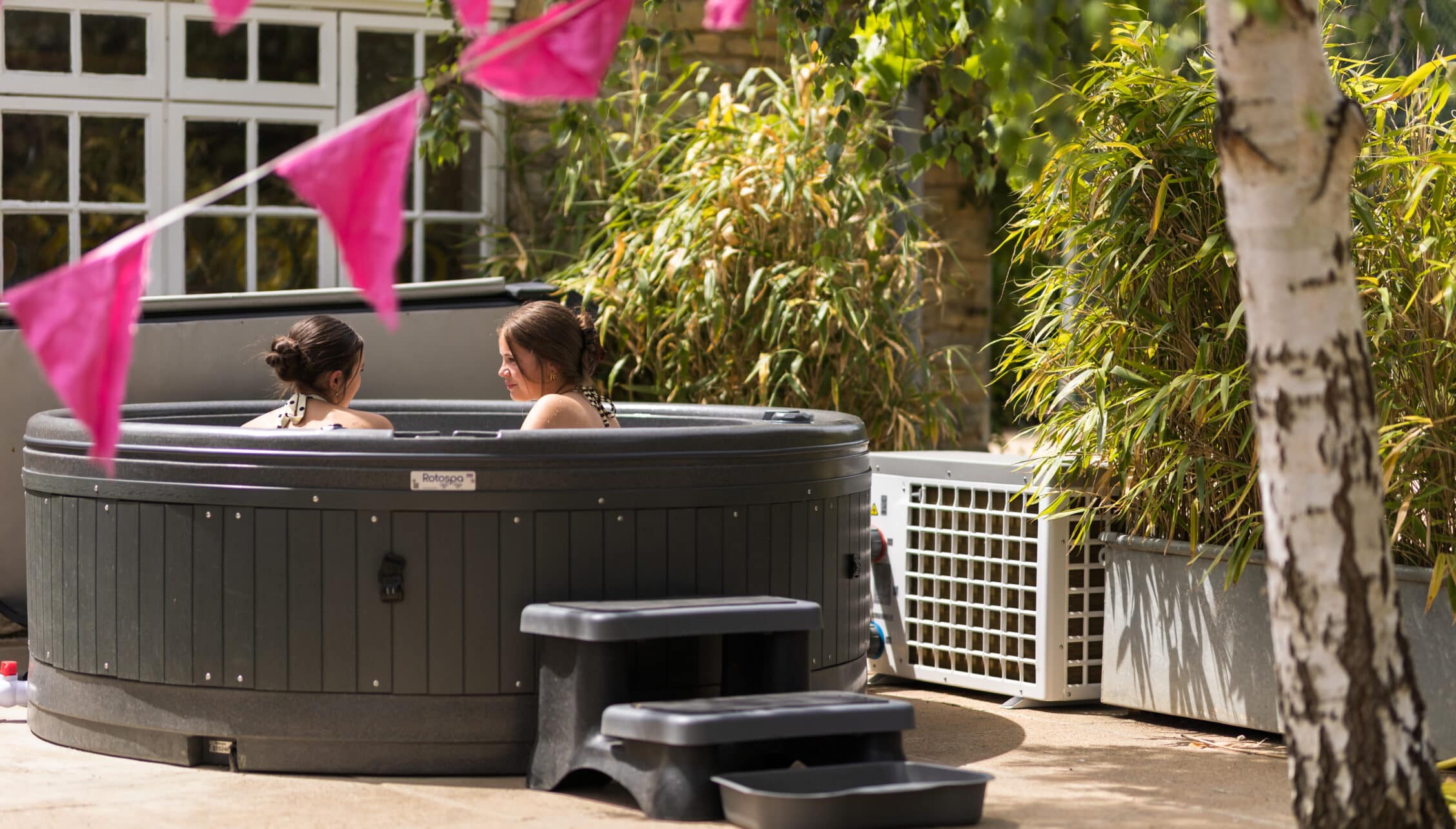 Courtyard Cottages, Gloucestershire – Two guests relaxing in hot tub with pink bunting and bamboo screening in courtyard garden.