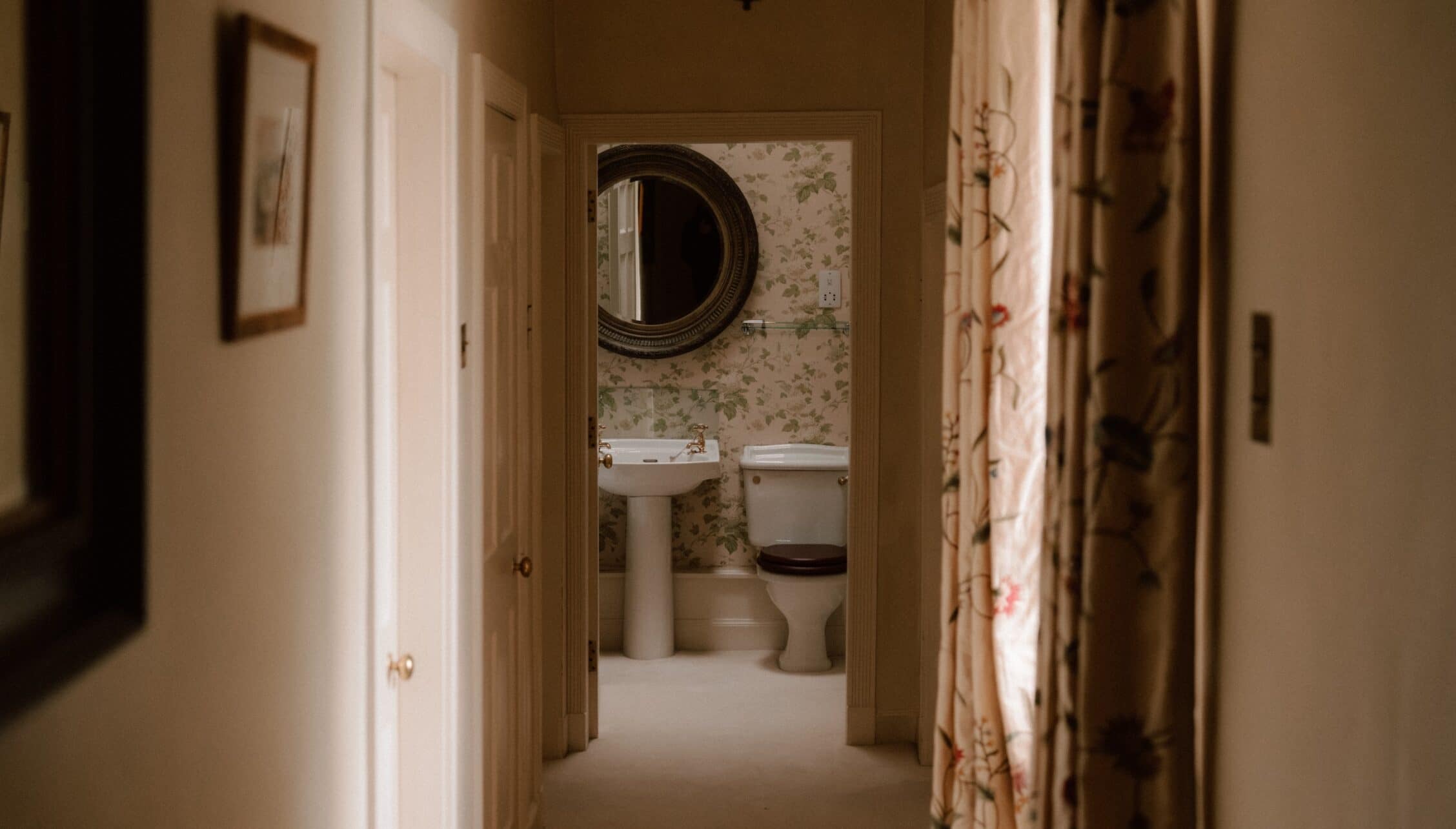 Ferniehirst Castle, East Lothian – Corridor view into bathroom with pedestal sink, toilet, and floral wallpaper.