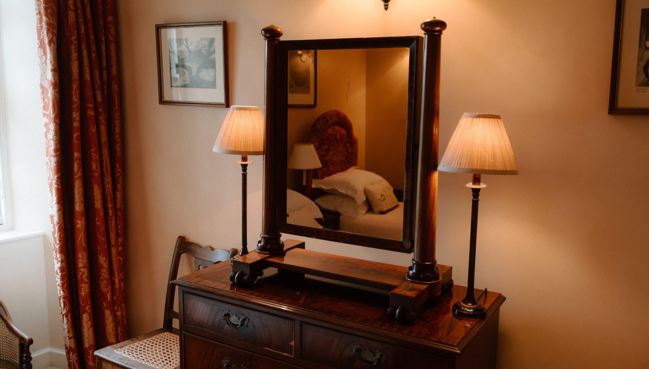 Ferniehirst Castle, East Lothian – Wooden dressing table with mirror and twin table lamps in bedroom with red curtains.