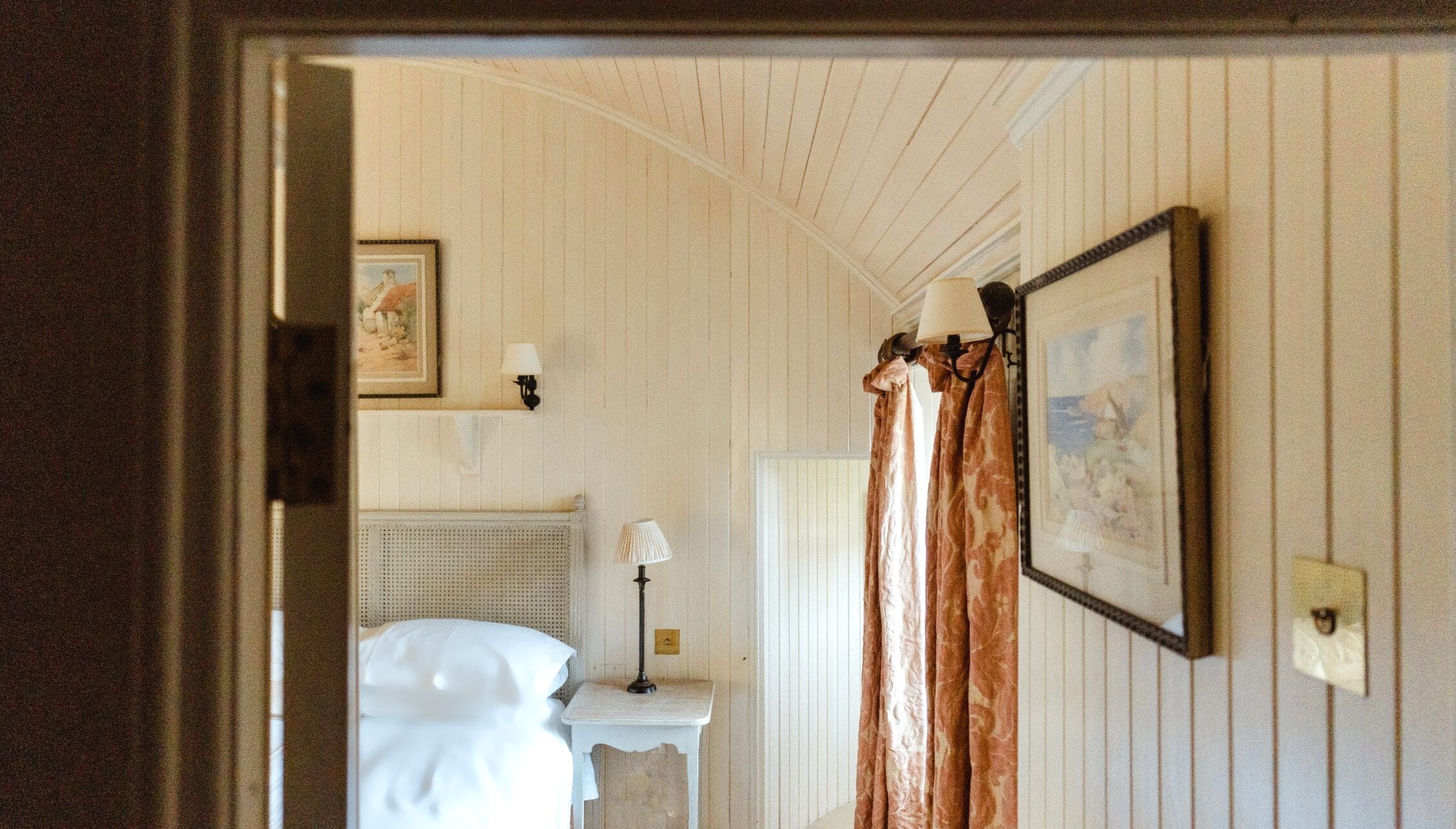 Ferniehirst Castle, East Lothian – Bedroom with cream shiplap walls, single bed with rattan headboard, and terracotta curtains.