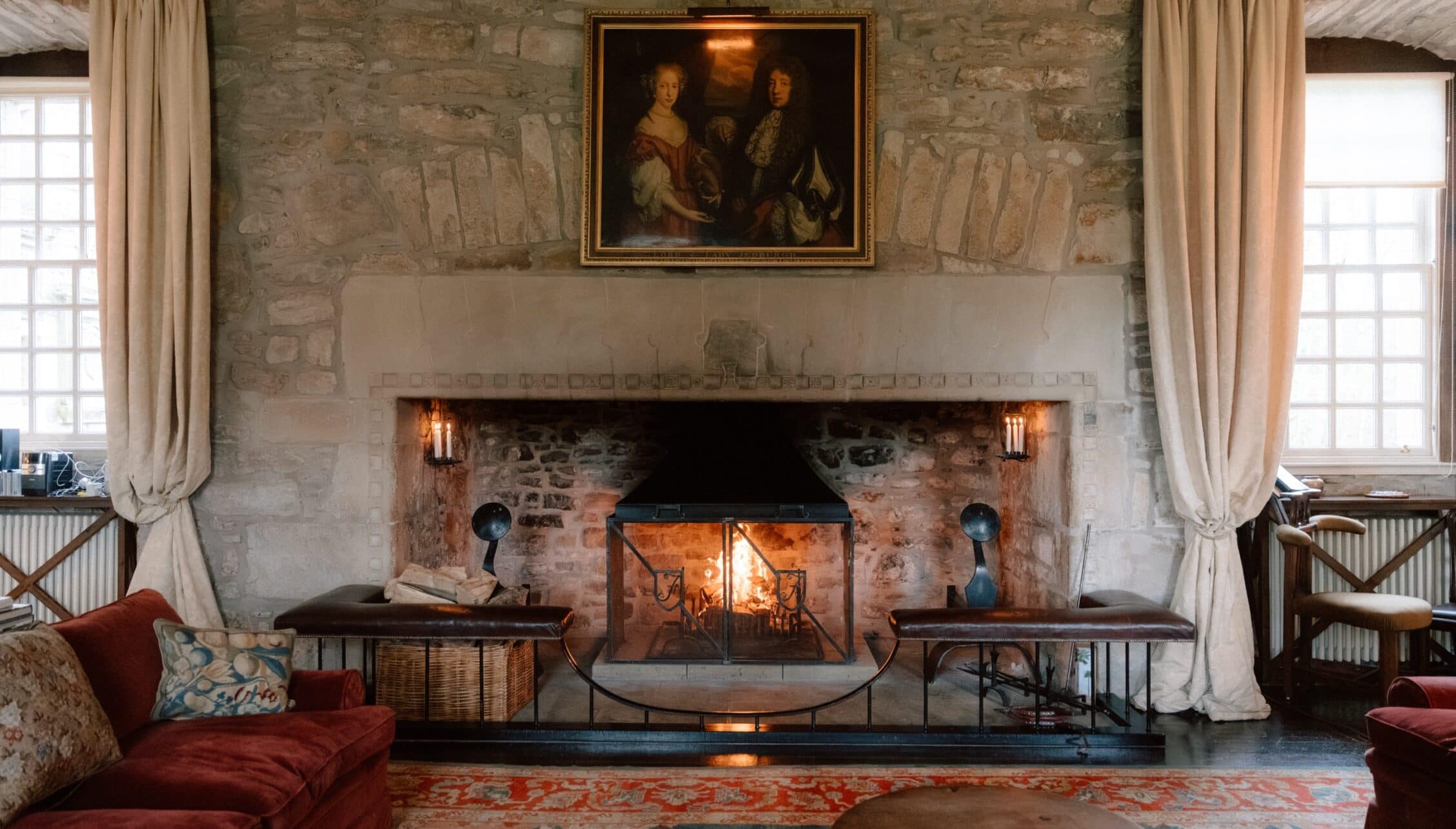 Ferniehirst Castle, East Lothian – Stone fireplace with lit fire, red velvet seating, and portrait above in large reception room.