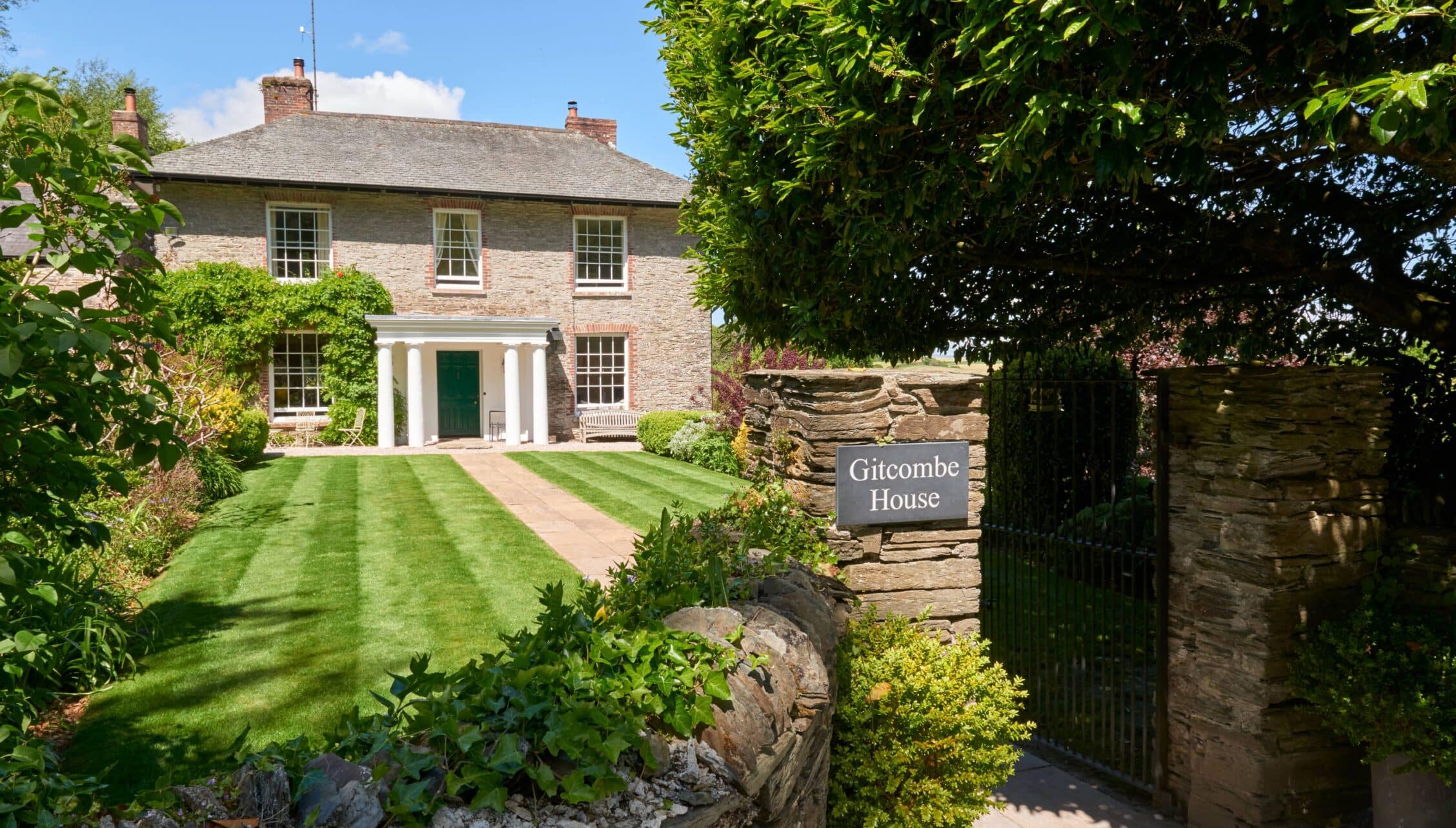 Gitcombe House, Devon – Brick country house with manicured lawn and formal entrance portico behind wooden gate.