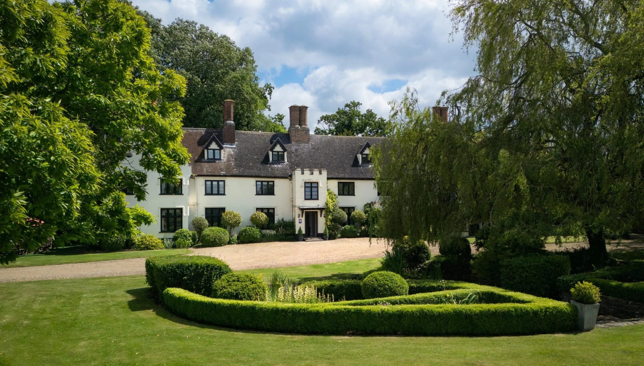 Hill House Norfolk, Norfolk – Cream-rendered country house with black gable windows, brick chimneys, gravel drive, and manicured grounds with topiary hedges.