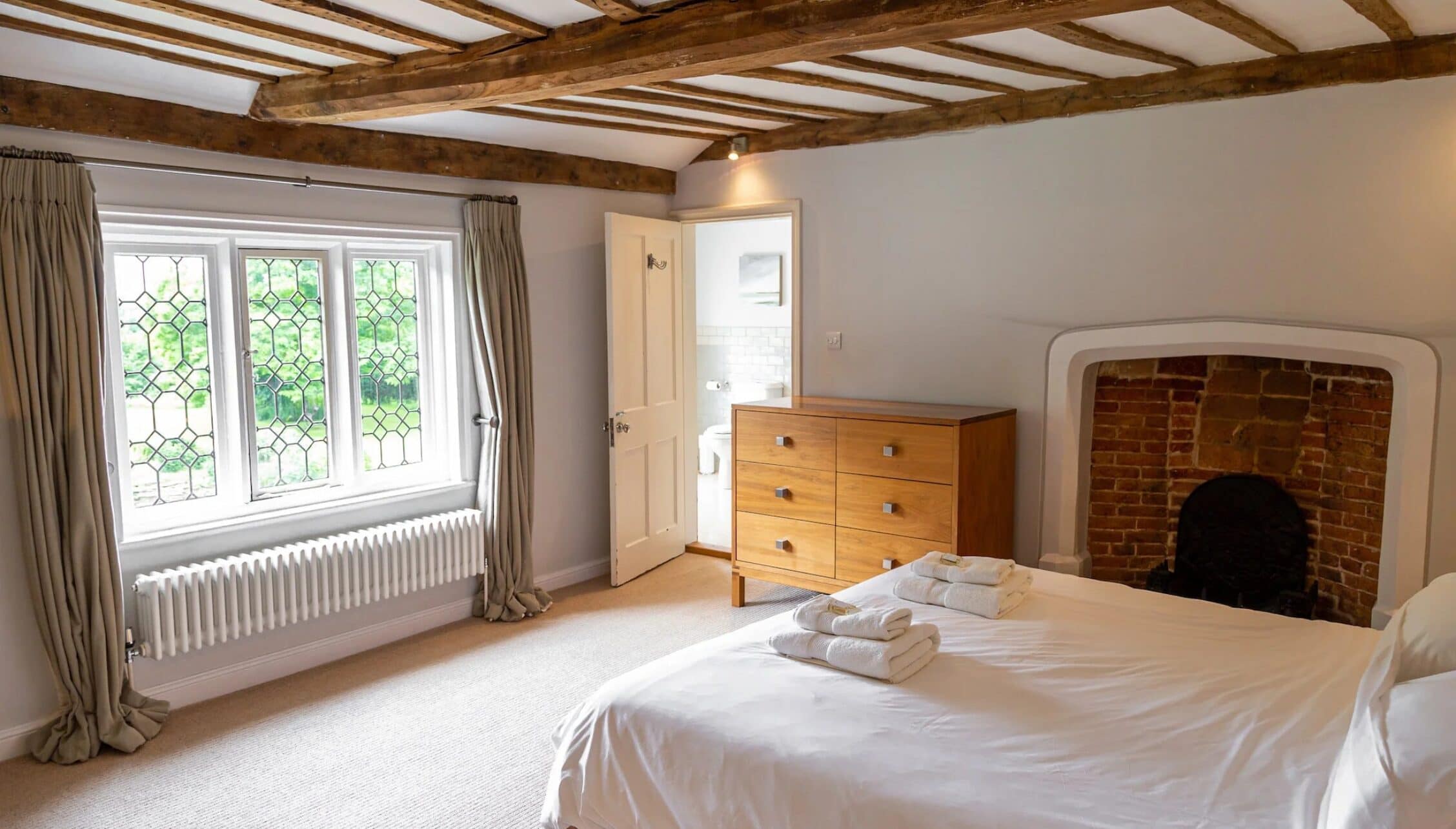 Hill House Norfolk, Norfolk – Spacious bedroom with exposed wooden beams, brick fireplace, and leaded windows.