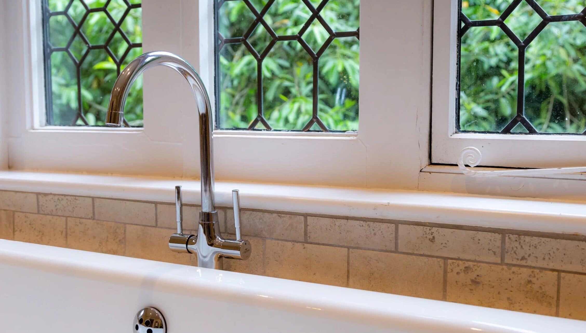 Hill House Norfolk, Norfolk – Chrome kitchen tap above white sink with leaded-glass windows and brick splashback.