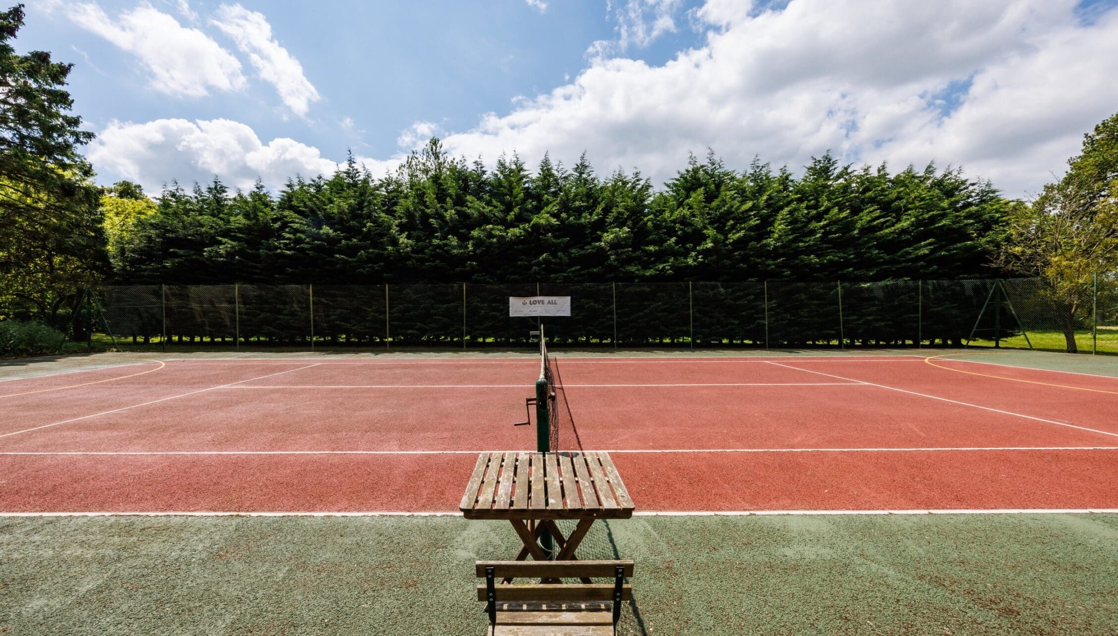 Hill House Norfolk, Norfolk – Red clay tennis court with net and wooden bench, surrounded by conifer windbreak.