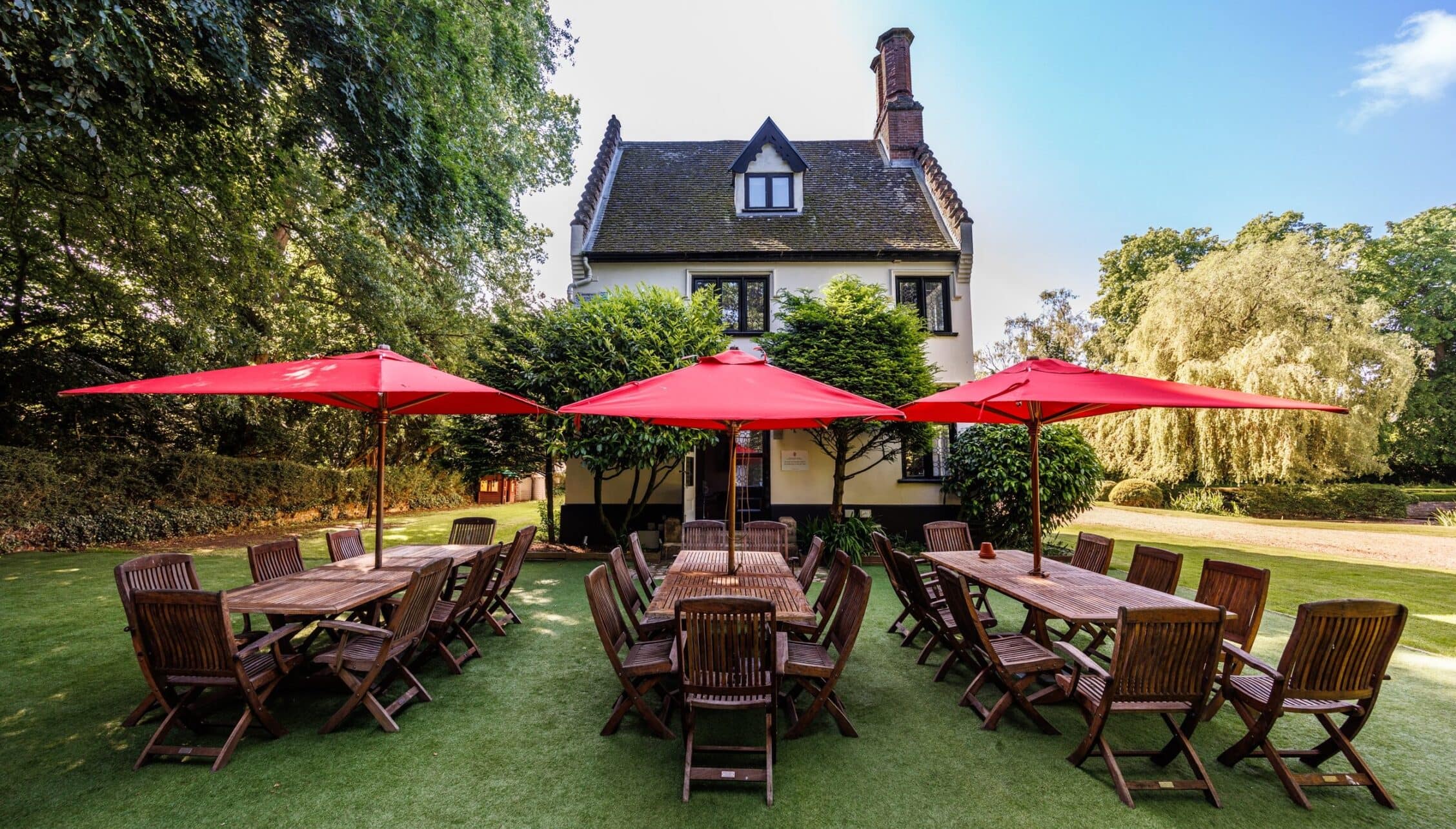 Hill House Norfolk, Norfolk – Lawned garden with wooden dining tables, red umbrellas and country house behind.