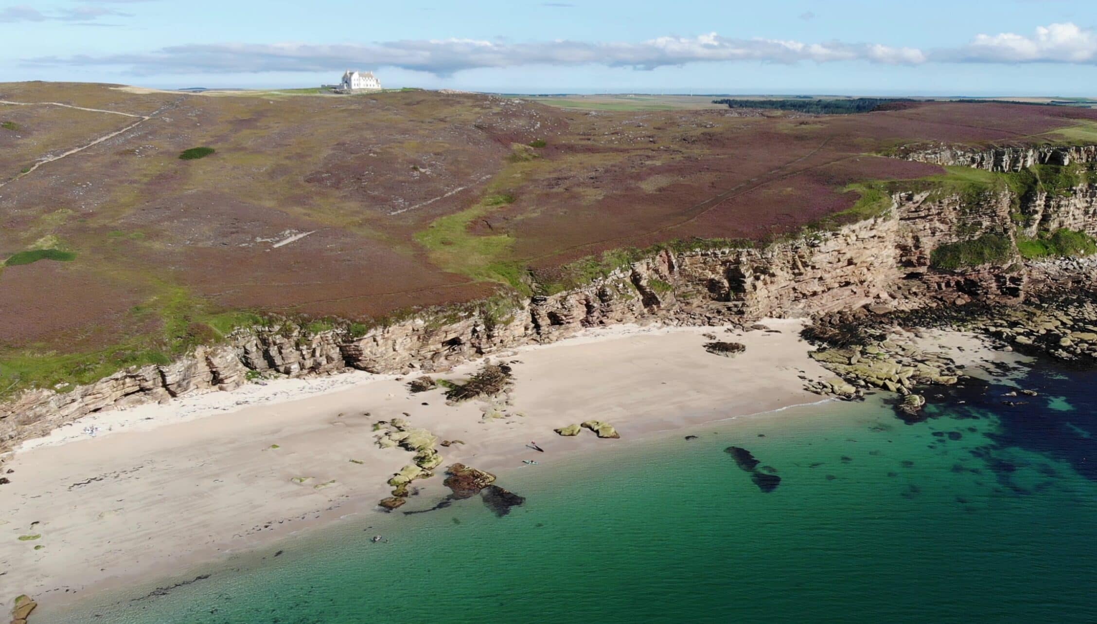 House of the Northern Gate, Highland – Coastal cliffs, sandy beach and turquoise water with distant white house on moorland.