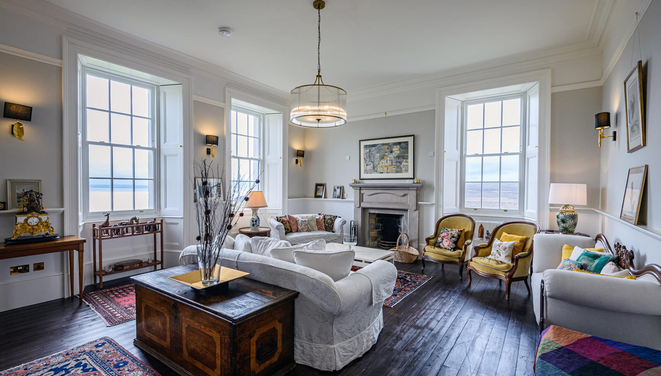House of the Northern Gate, Highland – Spacious sea-view sitting room with fireplace, sashed windows and mustard-yellow armchairs.