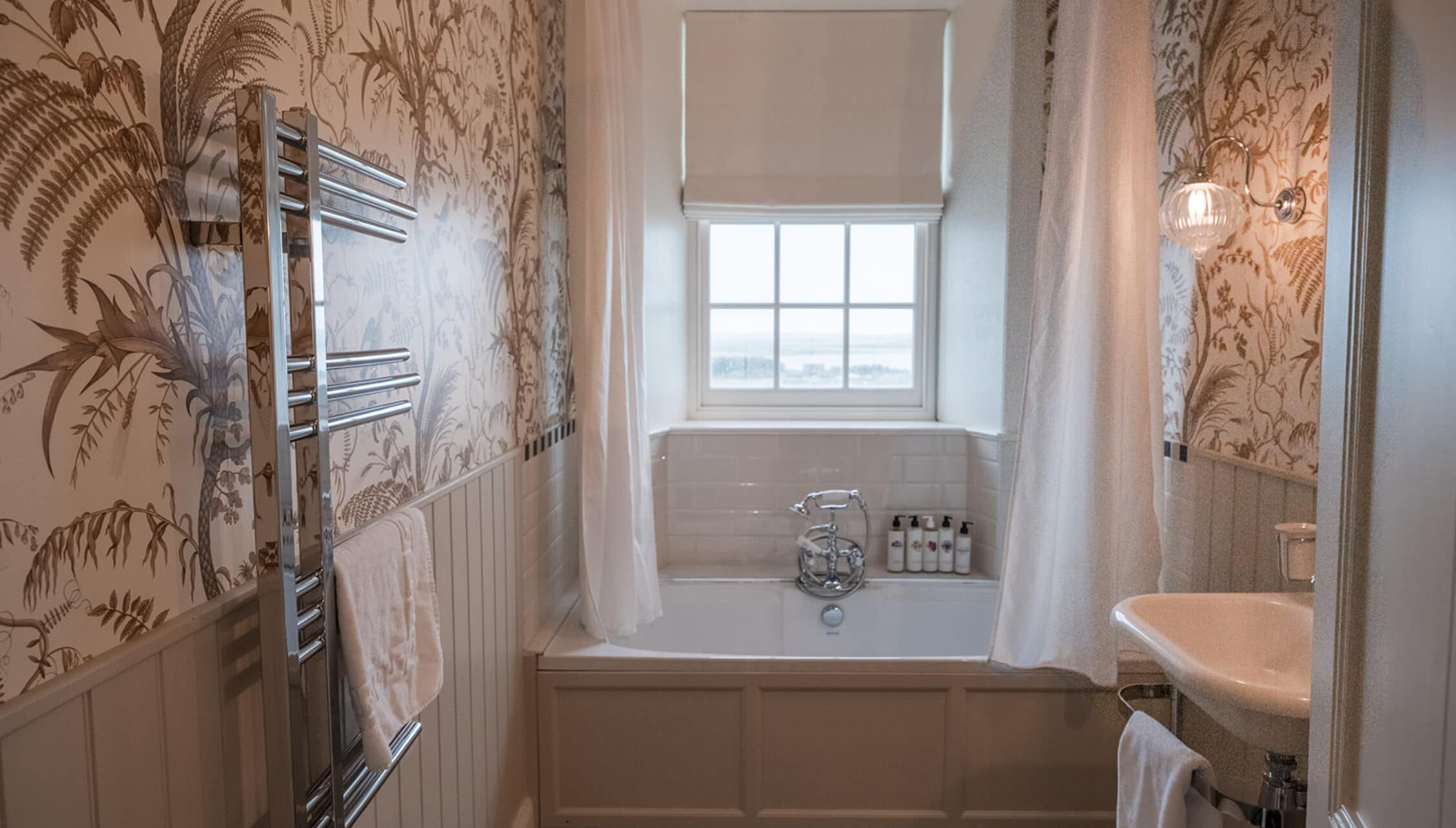 House of the Northern Gate, Highland – Bathroom with botanical wallpaper, heated towel rail, and bay window overlooking countryside.