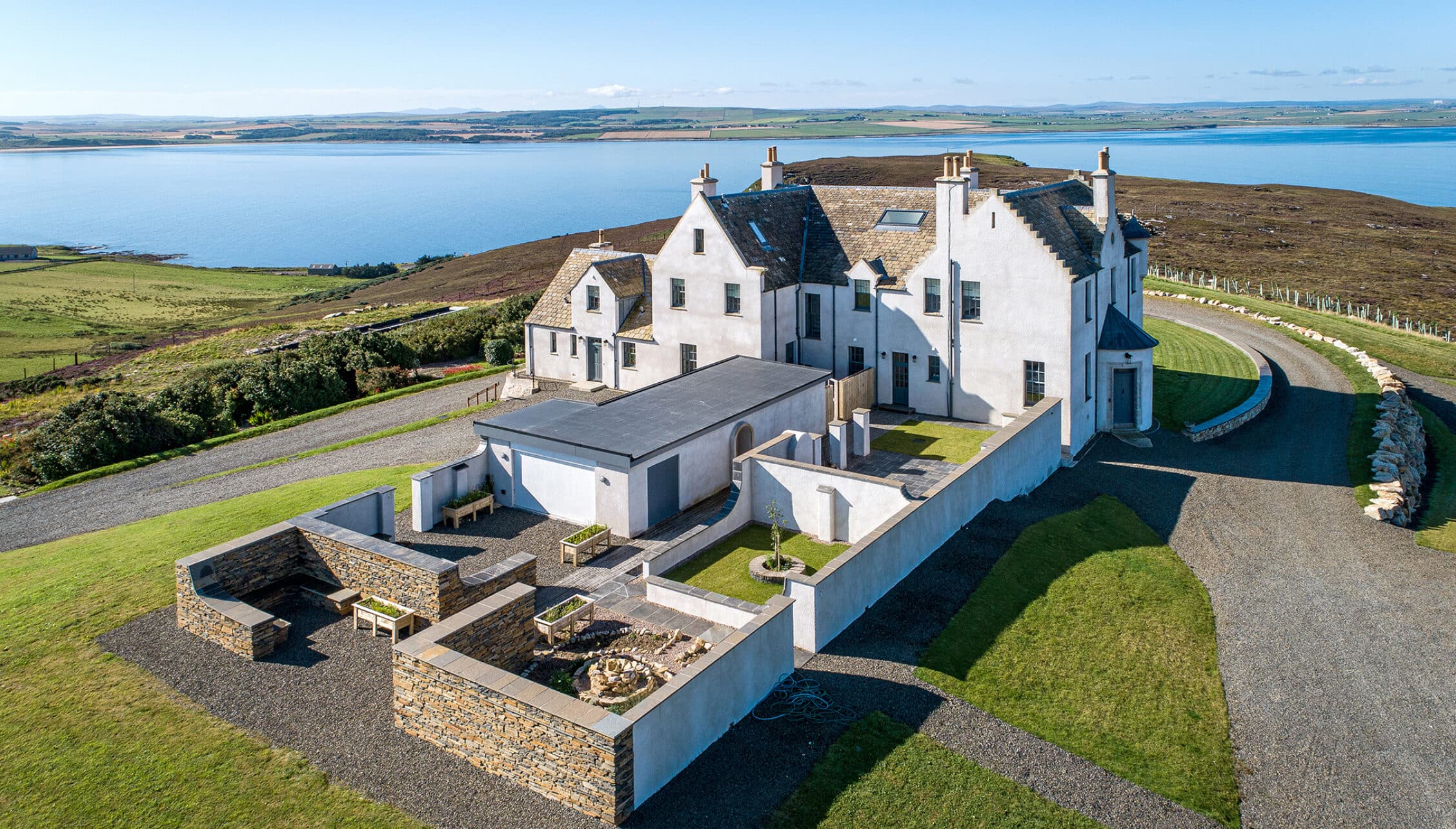 House of the Northern Gate, Highland – Modern white-rendered house with slate roof overlooking sea and mountains, surrounded by landscaped grounds with stone.