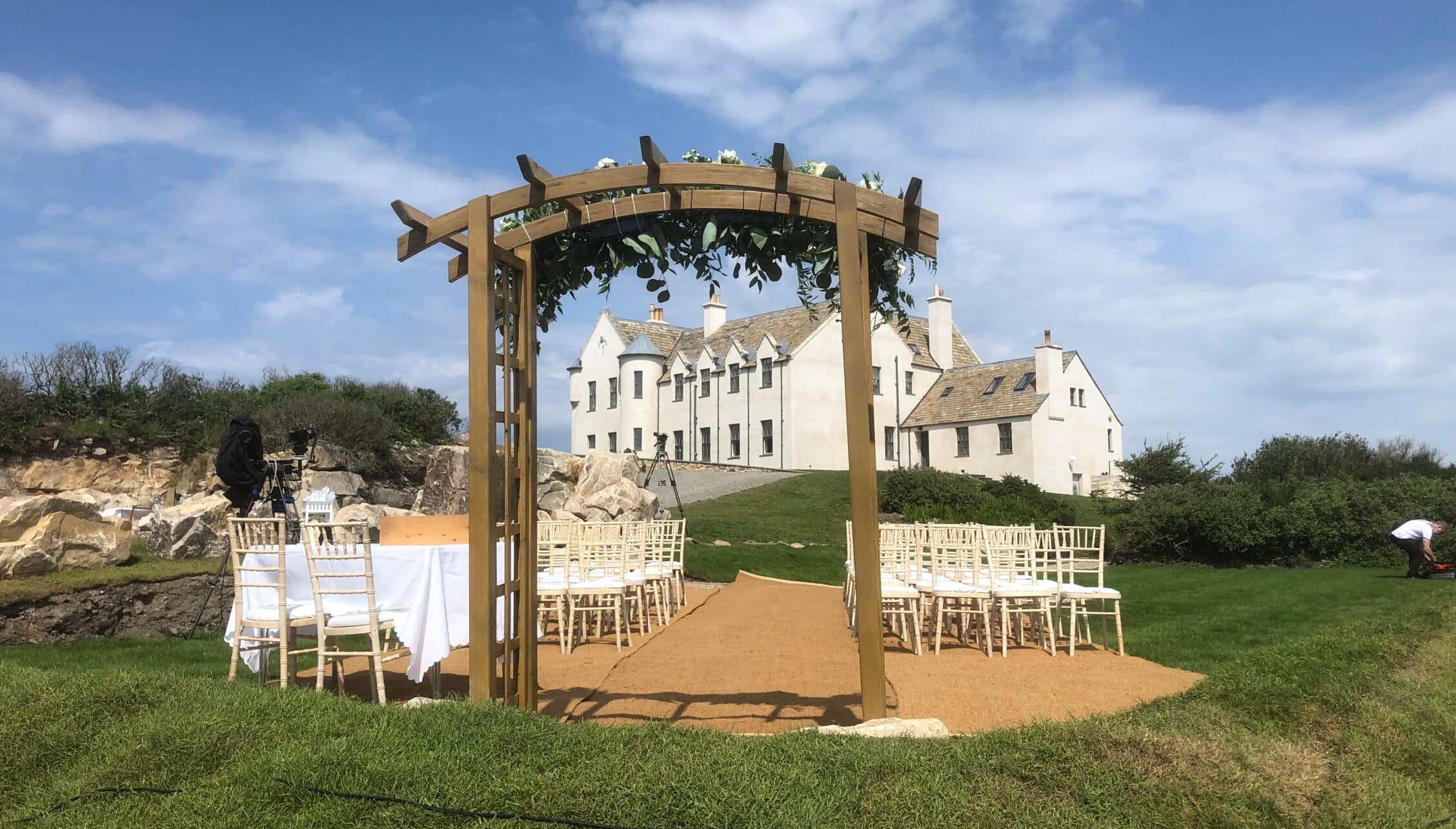 House of the Northern Gate, Highland – Wooden pergola with climbing foliage framing cream stone country house and seated guests.