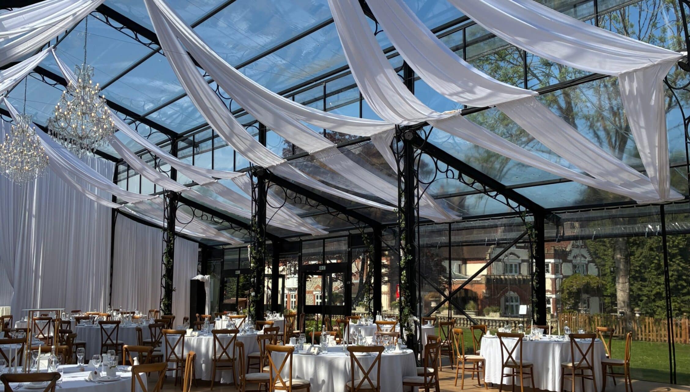 Kingswood Manor, Surrey – Glass pavilion with draped white fabric ceiling, wooden cross-back chairs, and crystal chandelier.