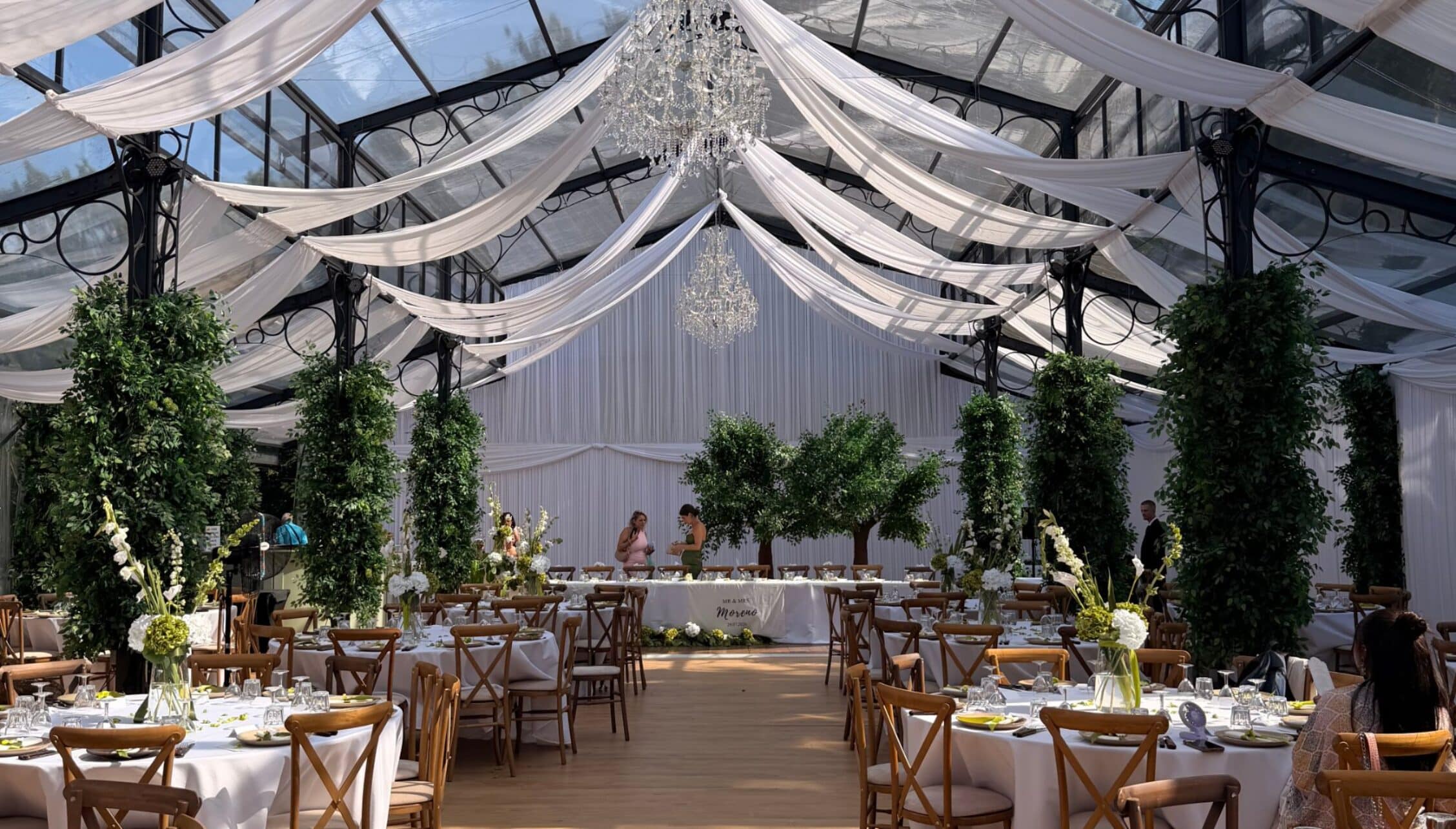 Kingswood Manor, Surrey – Marquee interior with draped sheer fabric ceiling, crystal chandelier and ivy-decorated wooden tables.
