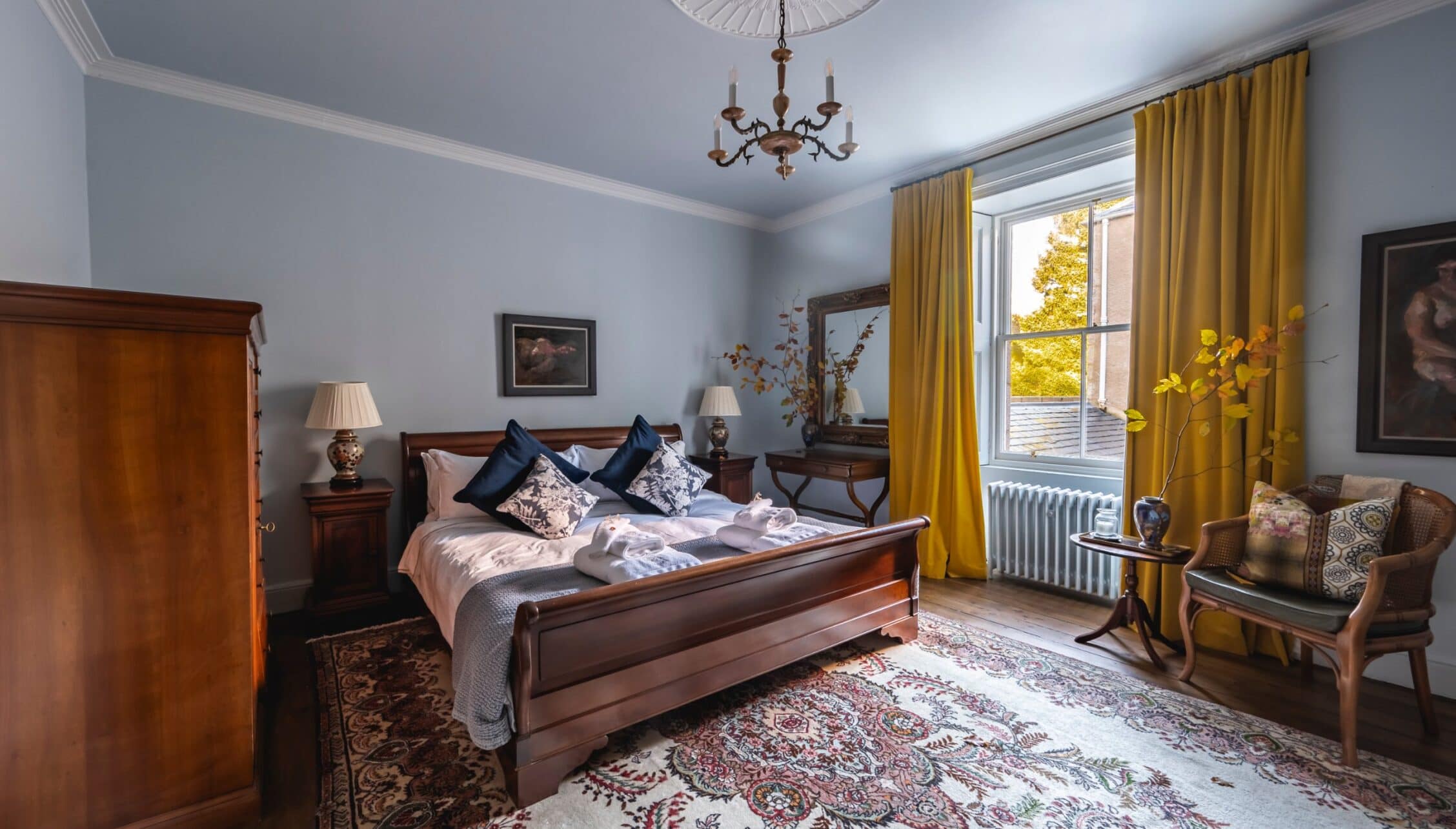 Manar House, Aberdeenshire – Master bedroom with mahogany bed, blue velvet cushions, and gold curtains overlooking autumn grounds.