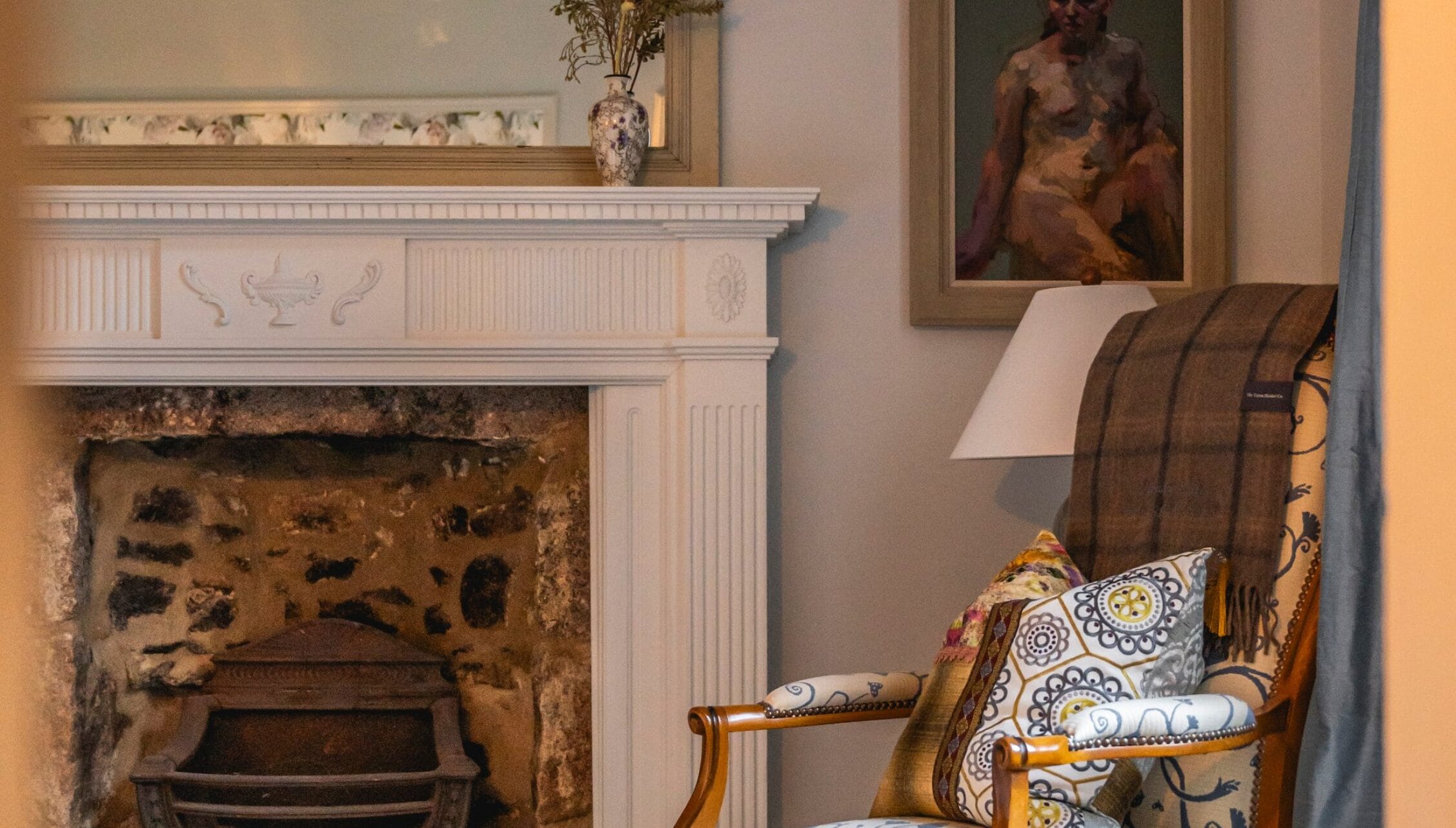 Manar House, Aberdeenshire – Cream painted fireplace with decorative moulding, wooden armchair with patterned cushions, and framed portrait.