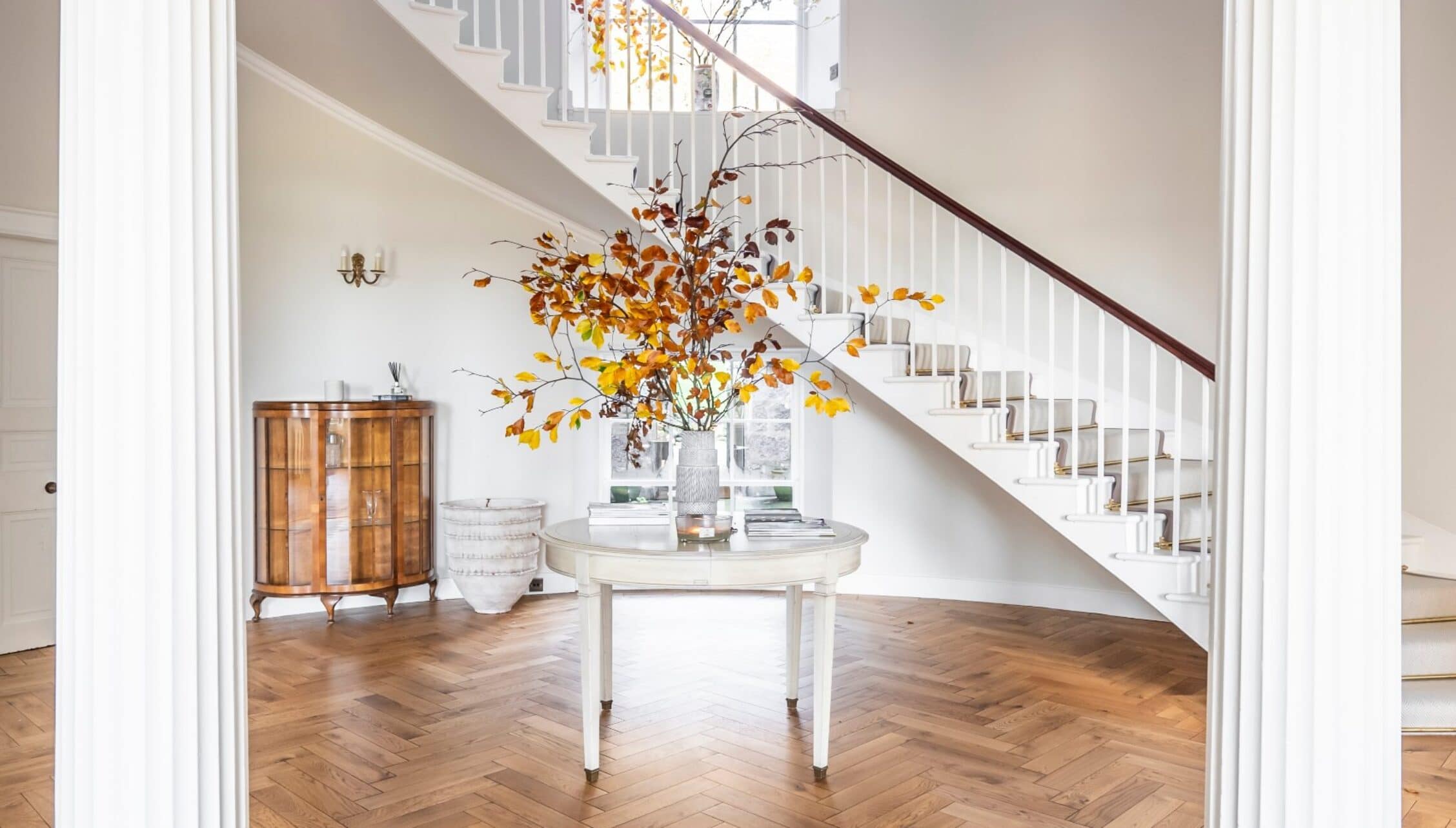 Manar House, Aberdeenshire – Entrance hall with curved staircase, autumn foliage arrangement and herringbone wooden flooring.